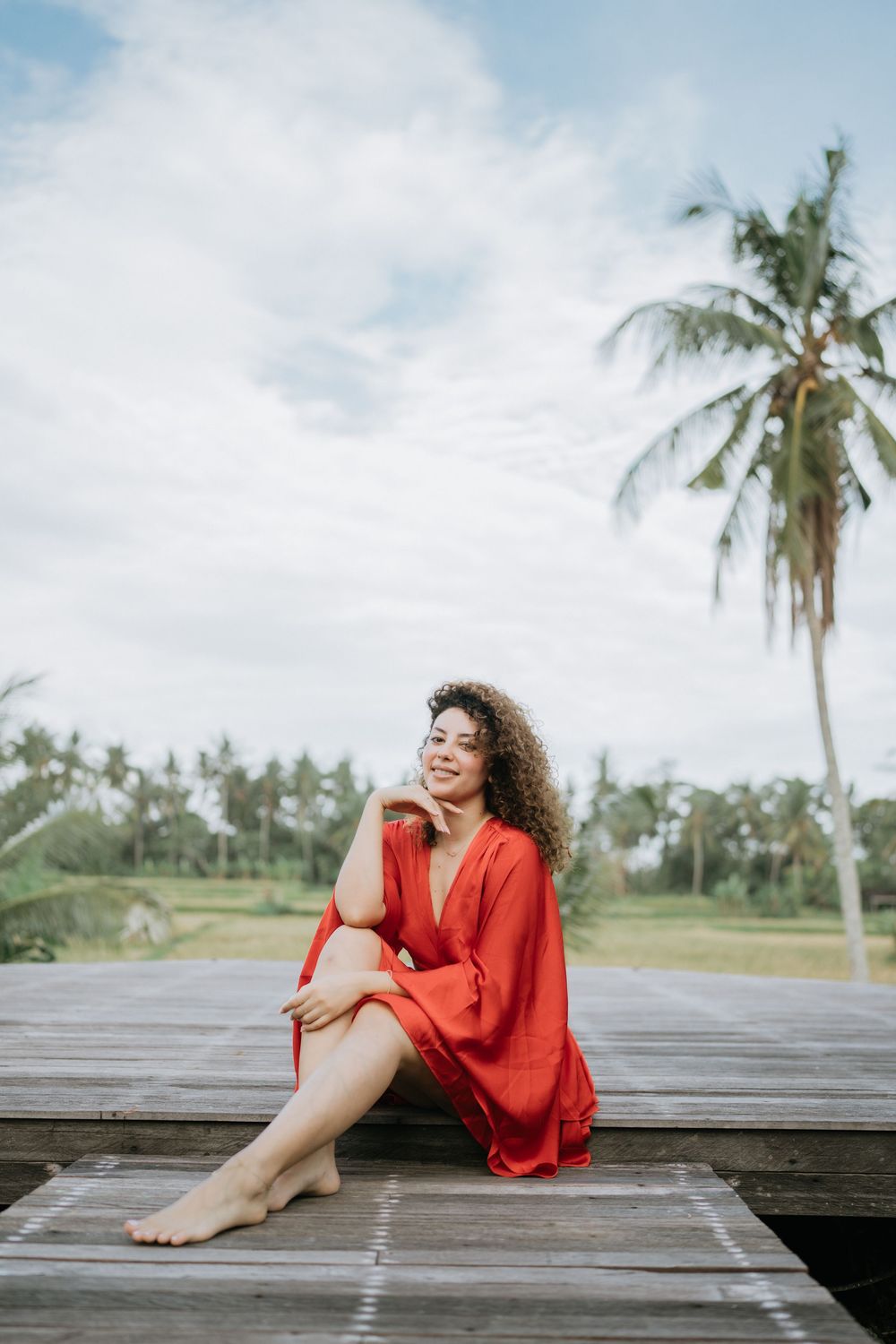 Person in red dress relaxing on wooden deck with palm trees against cloudy sky.