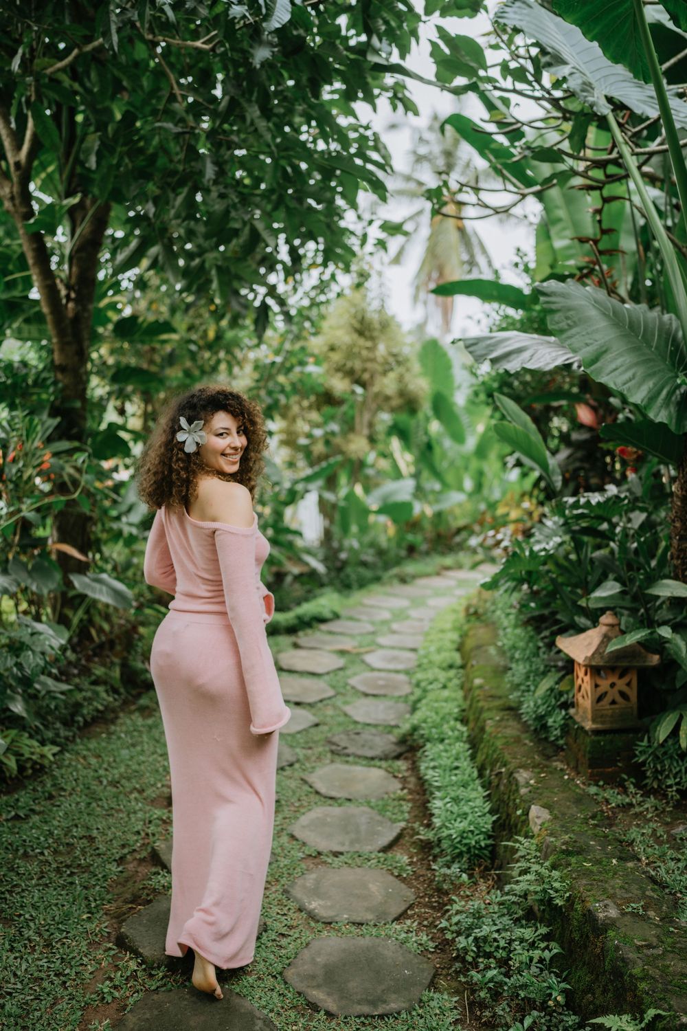 Person in pink dress walking along a stone path surrounded by lush tropical garden foliage.