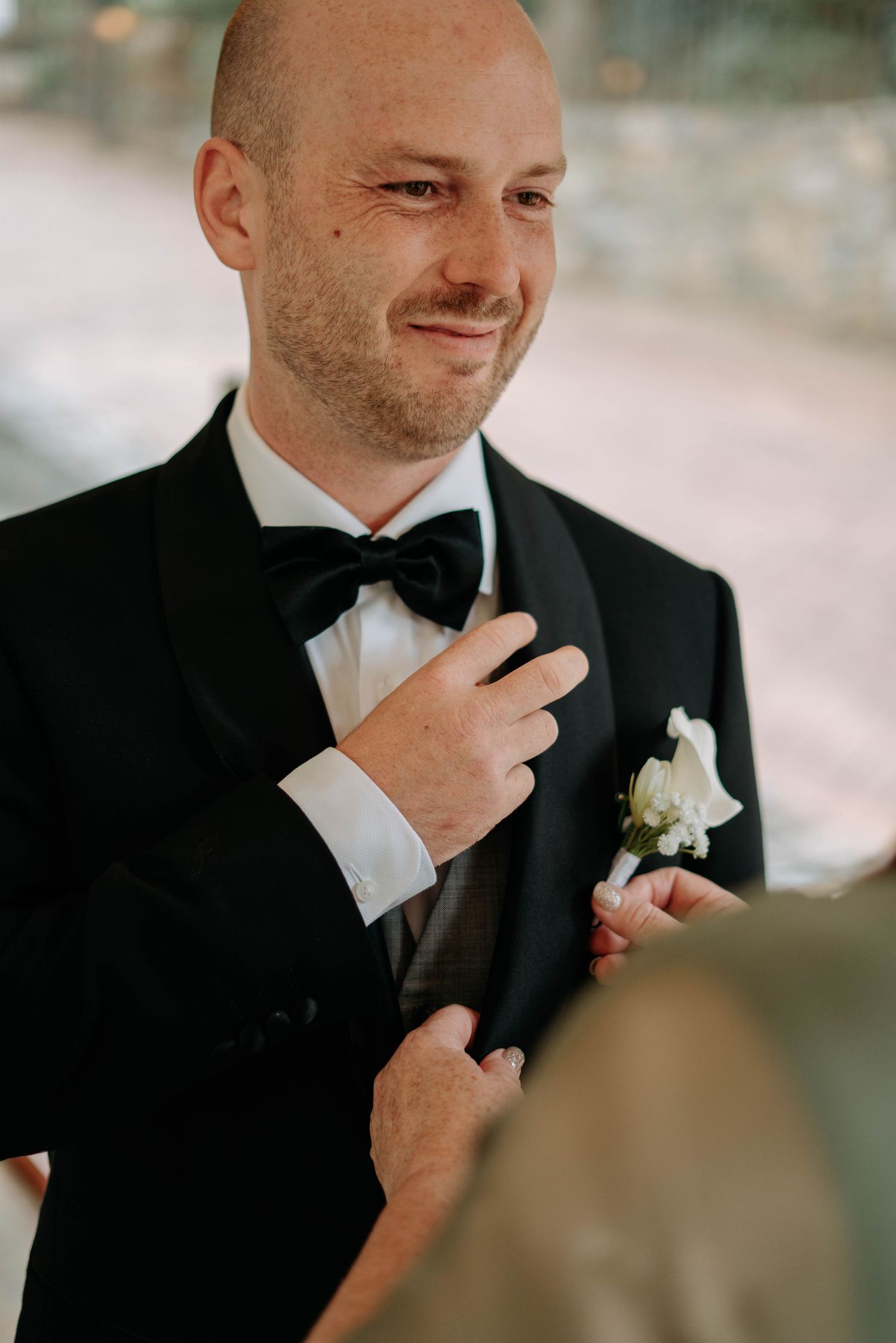 A groom in a black tuxedo adjusts his white boutonniere while getting ready for his wedding ceremony.