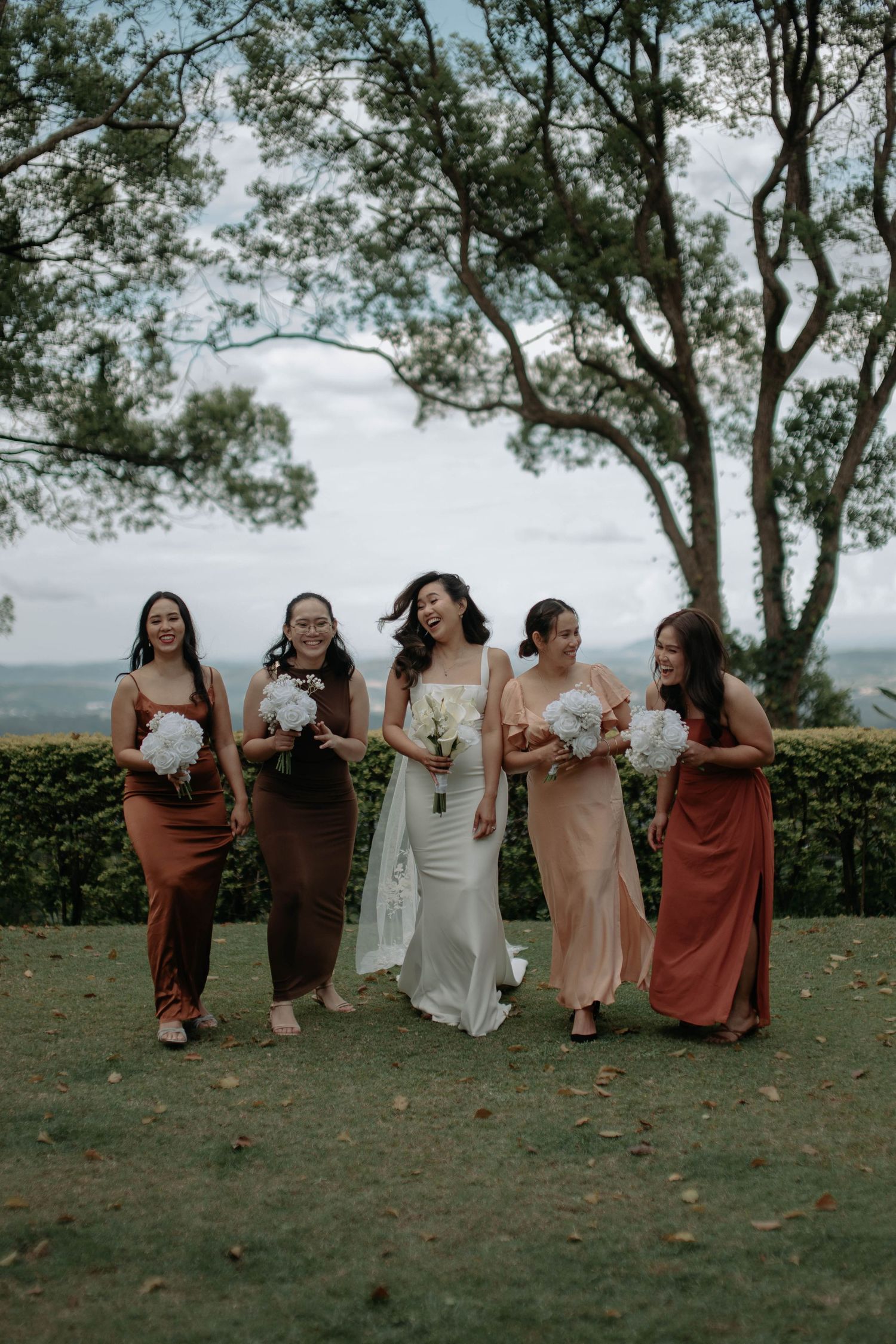 A group of friends in coordinated rust and cream colored dresses sit together on a grassy hillside overlooking a scenic view.