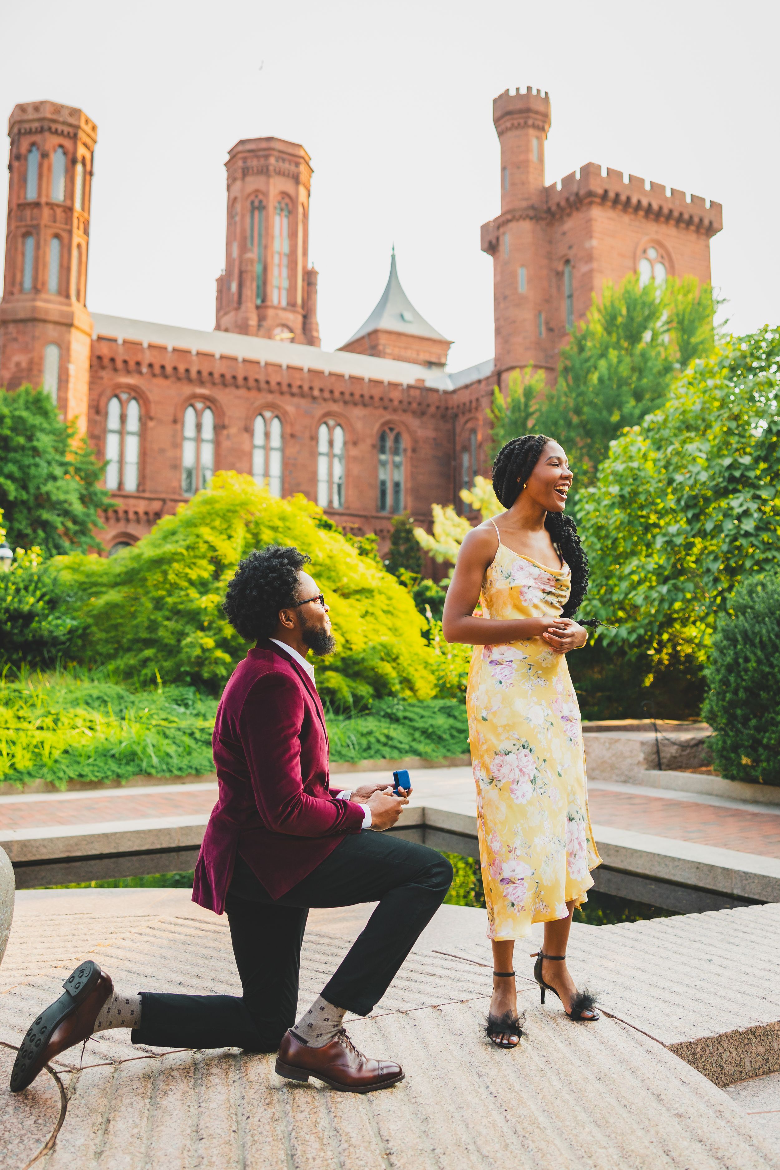 A joyful moment captured during a marriage proposal in front of the Smithsonian Castle with vibrant greenery.