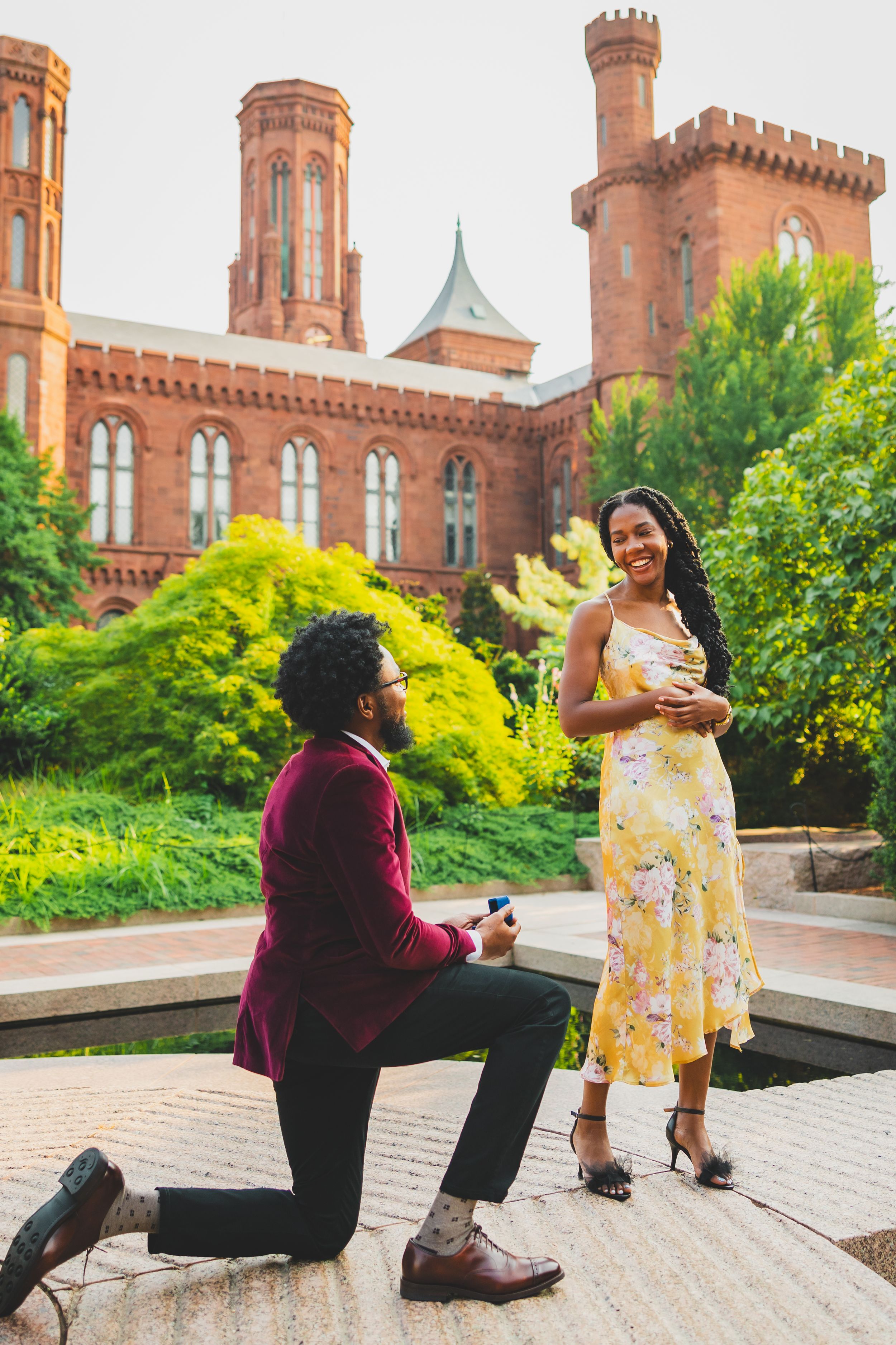 A proposal scene unfolds on a brick walkway in front of a historic castle-like building with red brick towers.
