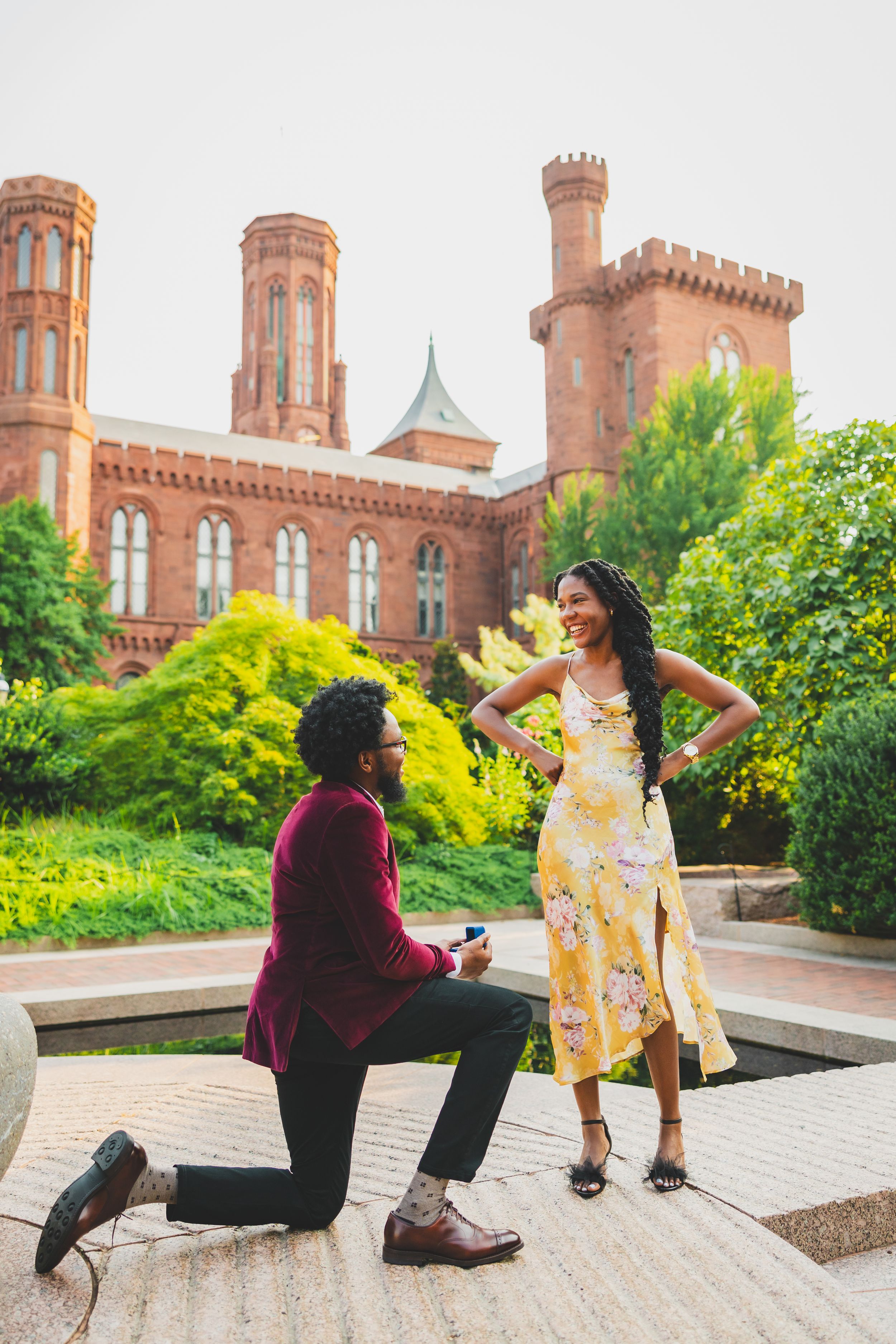 A romantic proposal takes place in front of a historic castle-like building with red brick towers and lush gardens.