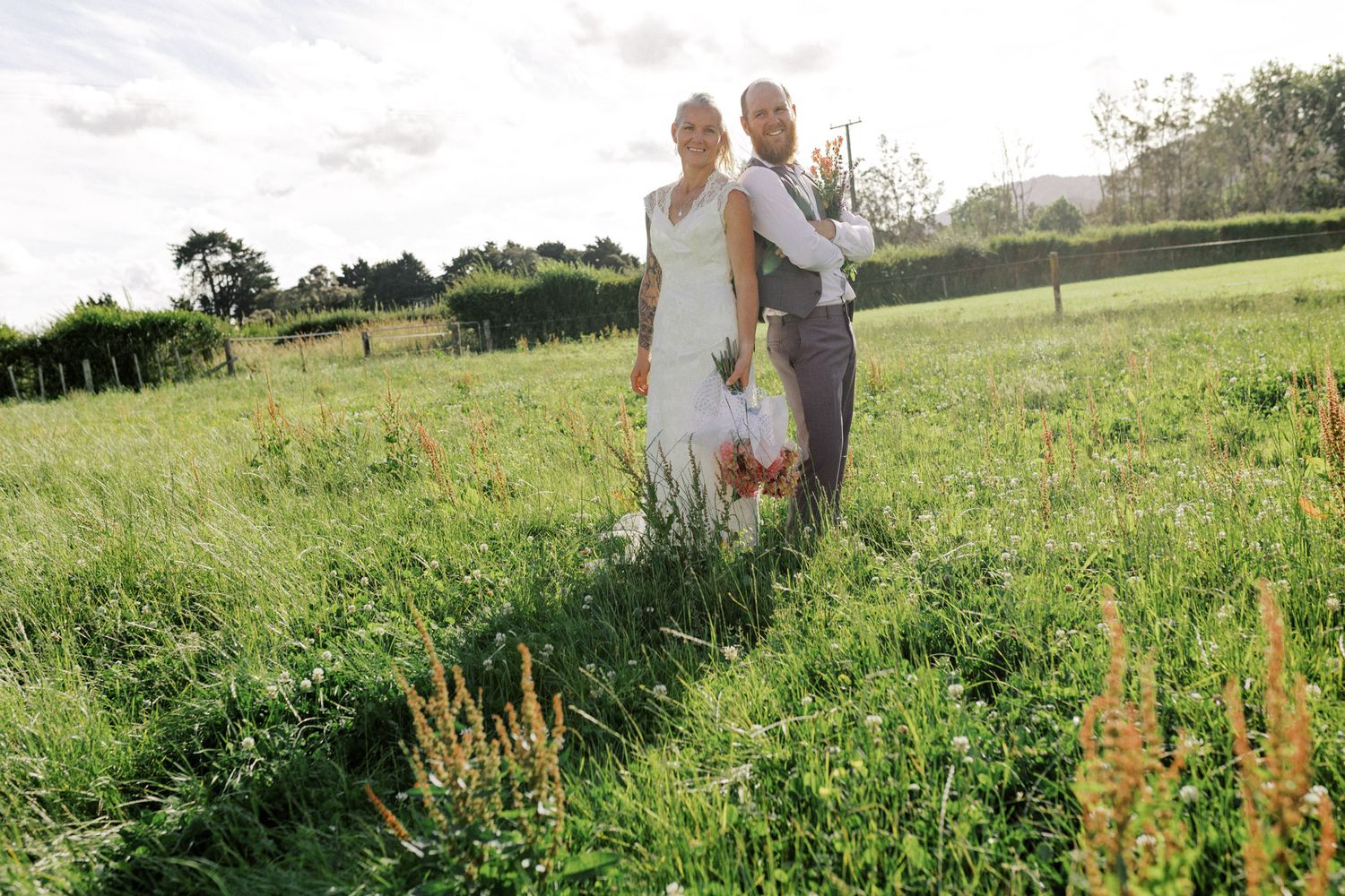 A couple walks together through a lush green meadow on what appears to be their wedding day.