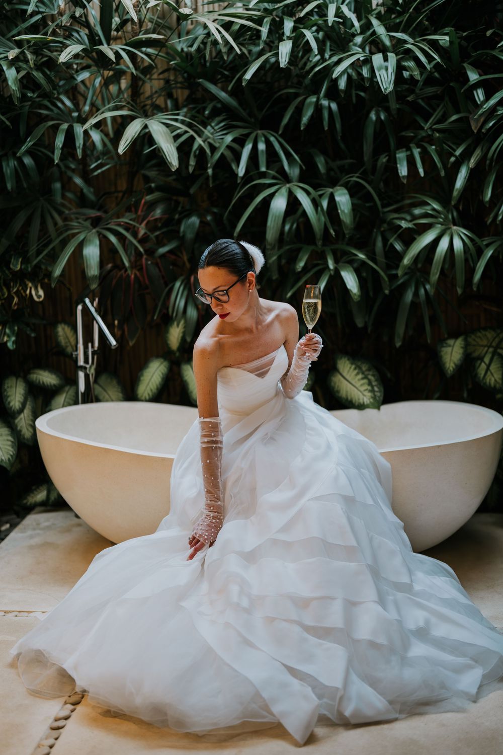 Artistic shots of a bride in glasses sitting by a freestanding bathtub surrounded by lush tropical foliage.