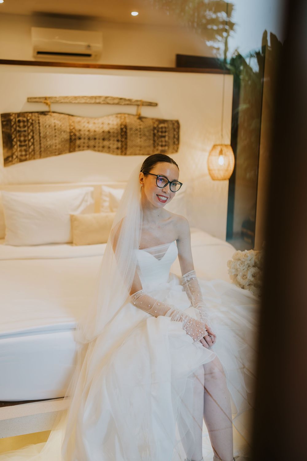 Interior shots of a bride in a flowing white dress seated on a plush bed in a modern room.
