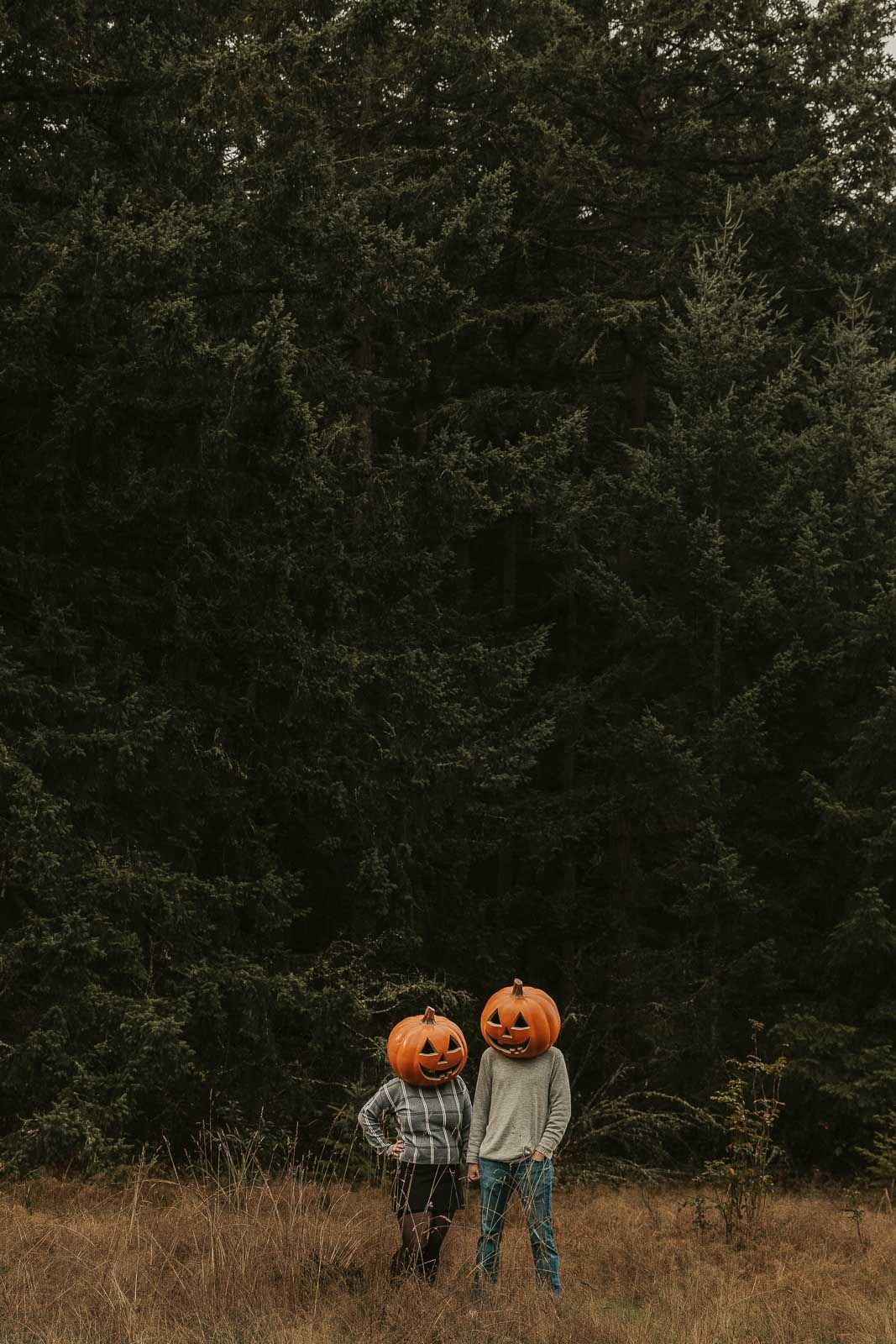 spooky Halloween couples photo shoot man and woman with pumpkin heads in open field with tall pine trees in background, spokane photography