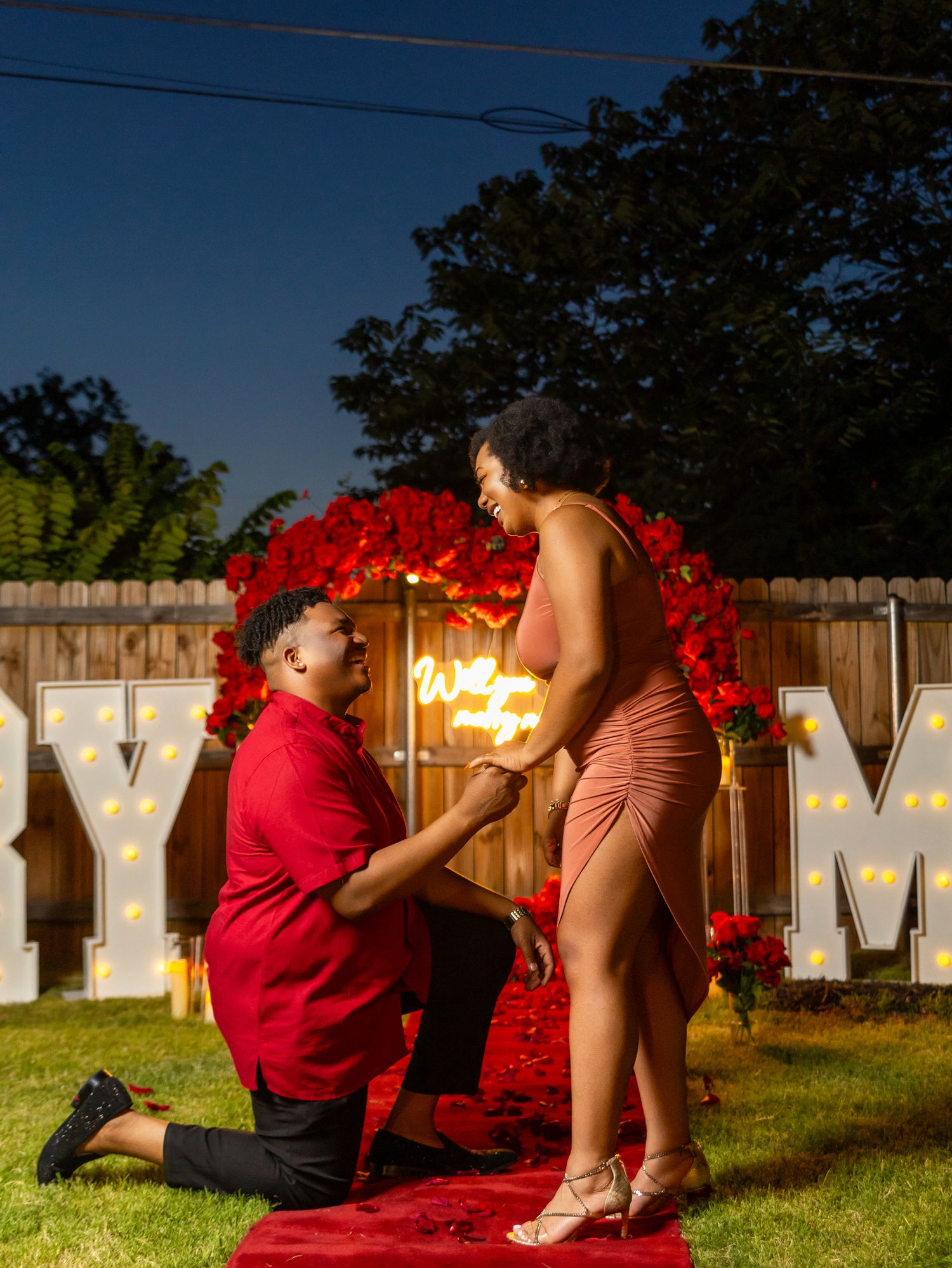 Romantic outdoor proposal scene with illuminated letters at night.