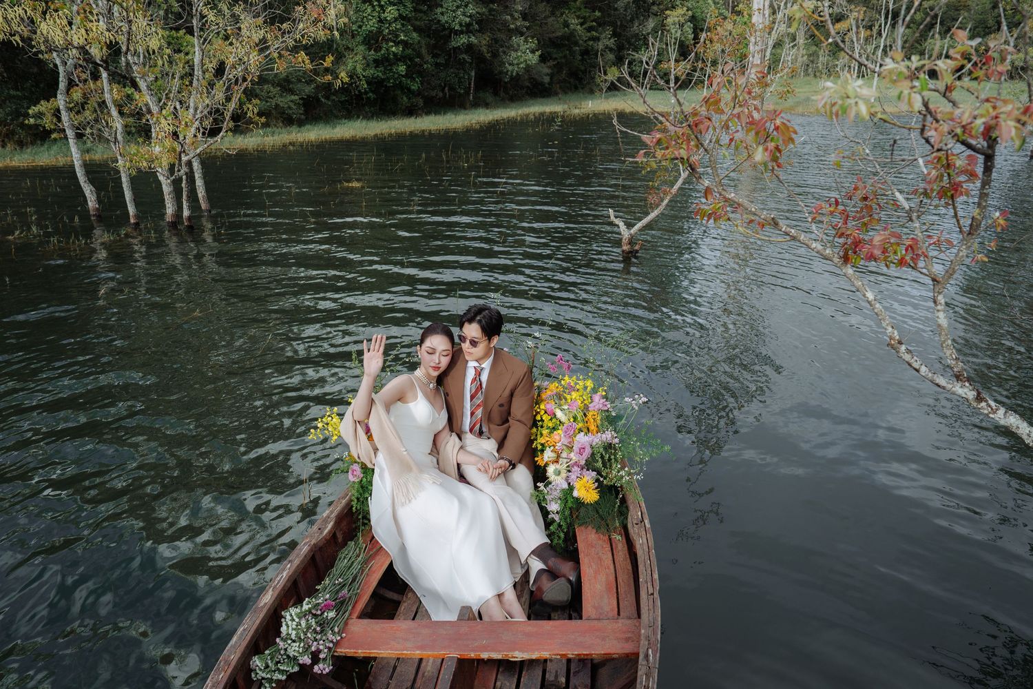 Wedding couple seated in a rustic wooden boat with flowers on a calm tree-lined lake, smiling warmly.