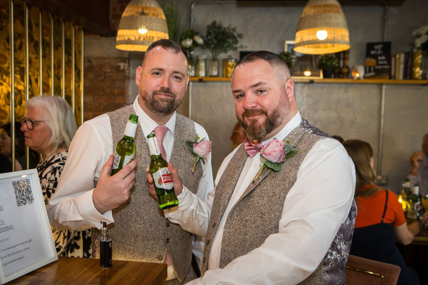 Two people in matching tweed vests toast with beer bottles at a casual wedding reception.