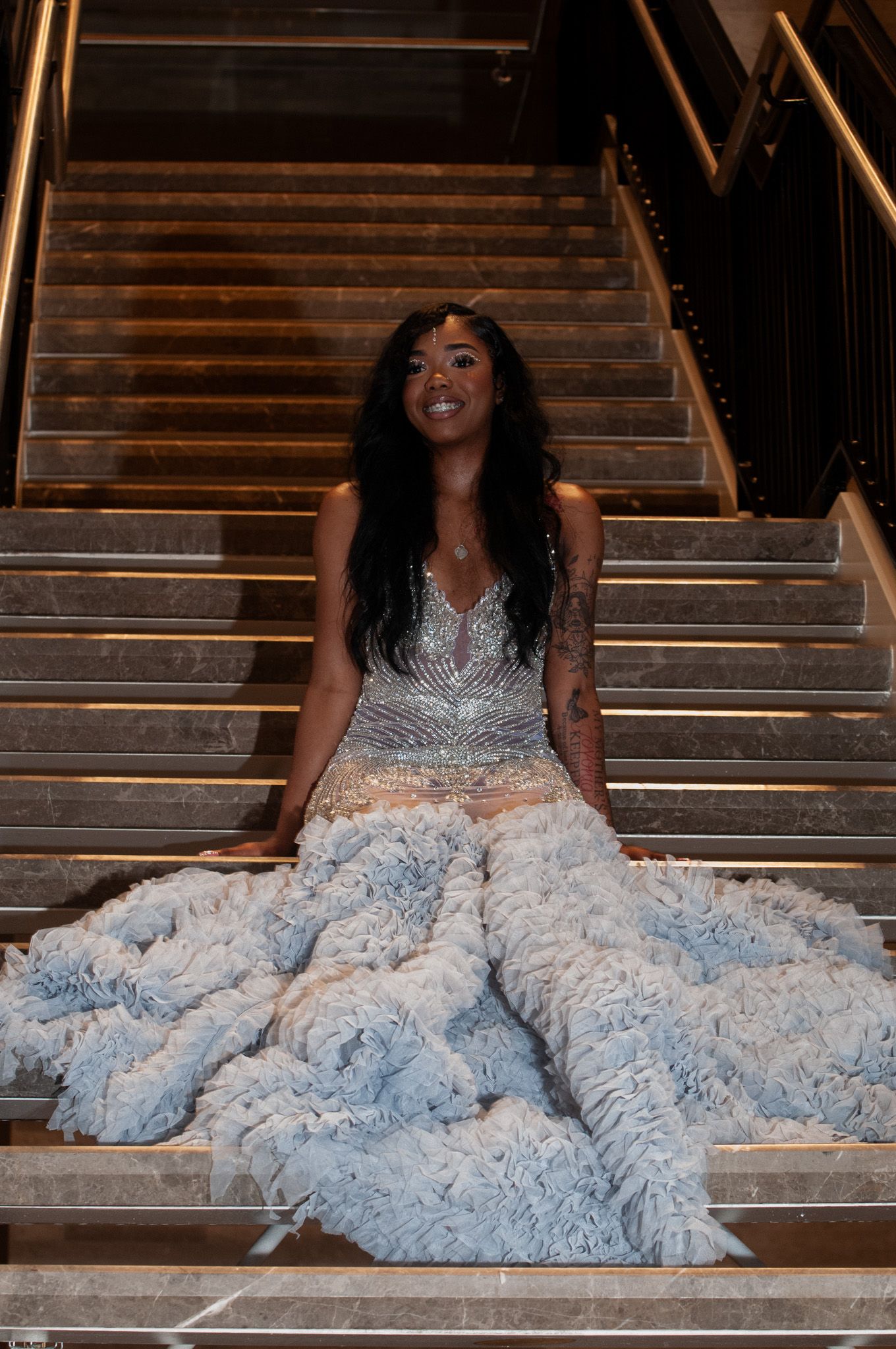 Woman in elegant light blue feathered gown sitting gracefully on grand staircase.