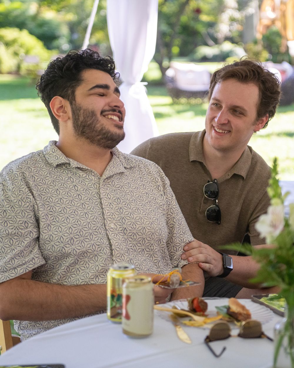 Two friends enjoying food and drinks at an outdoor table under white tent.