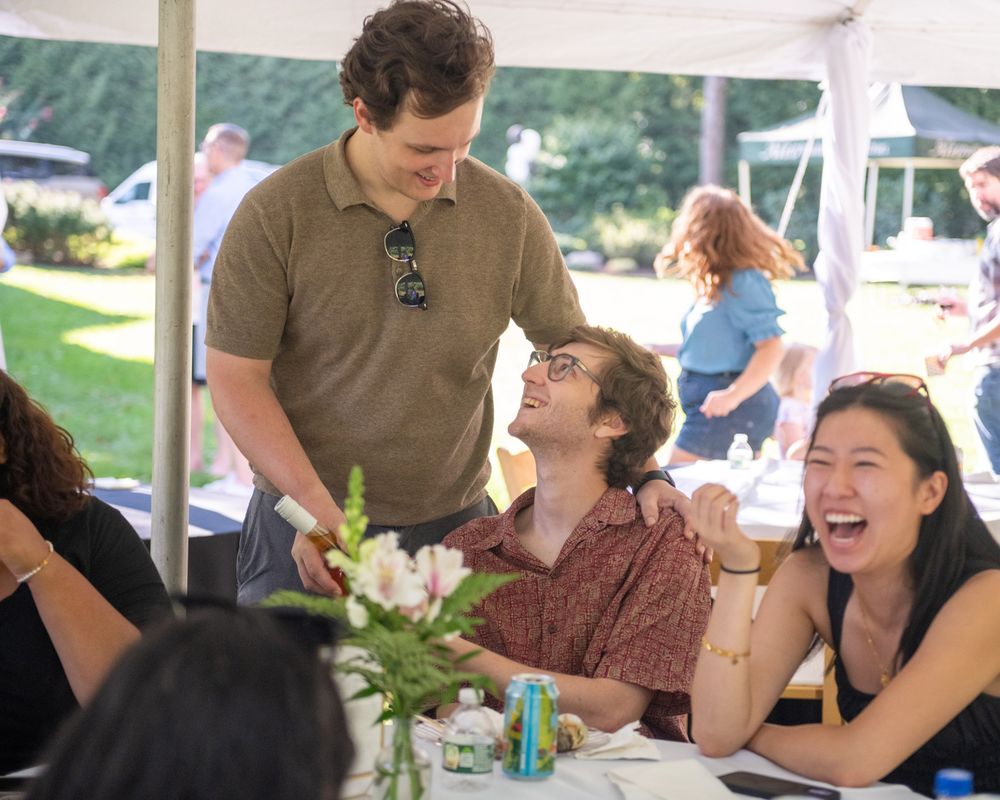 People laughing and socializing under a white tent at an outdoor event.
