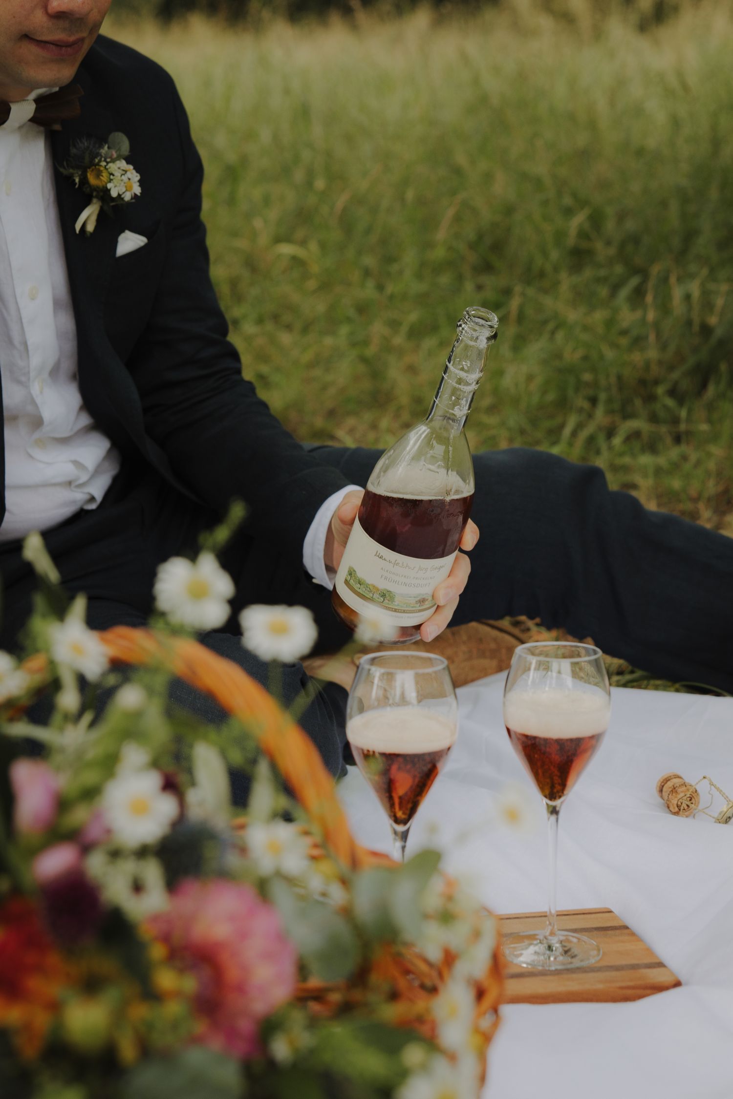 A couple enjoys wine and appetizers on a white blanket at an outdoor picnic with colorful flowers nearby.