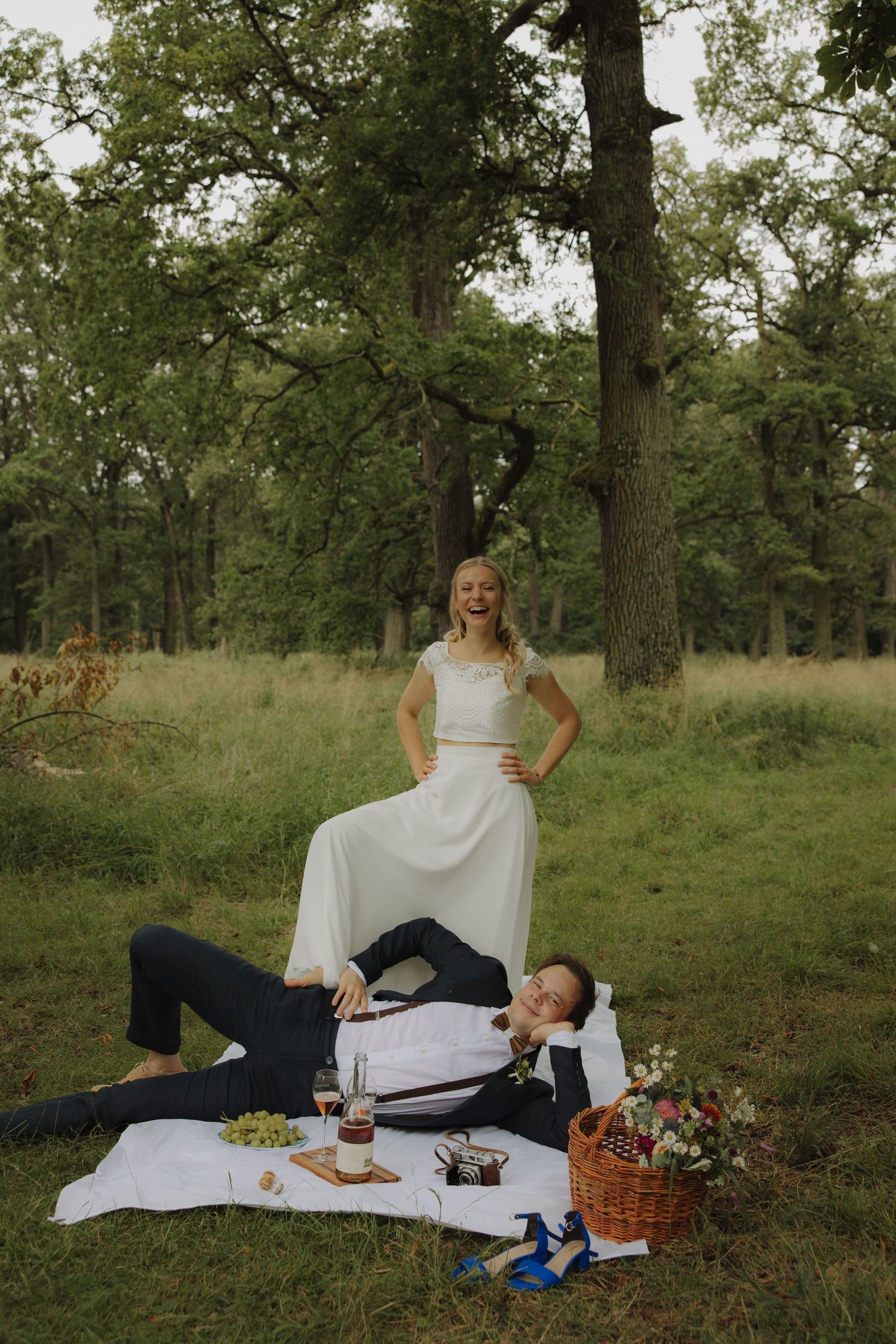 Couple enjoys a casual wedding picnic with wine and snacks on a white blanket in a wooded area.