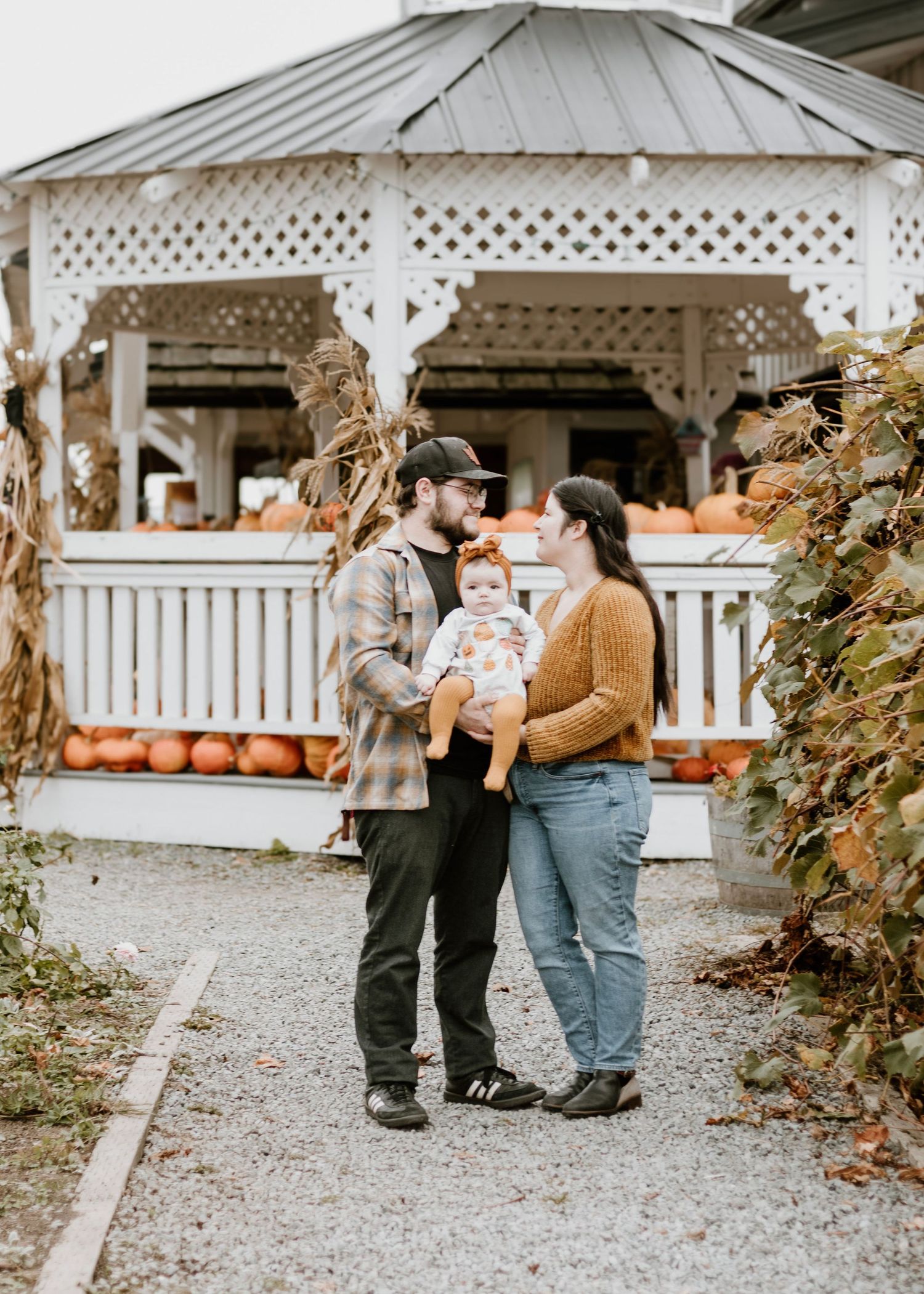 Family standing by white gazebo surrounded by fall pumpkins in autumn setting.