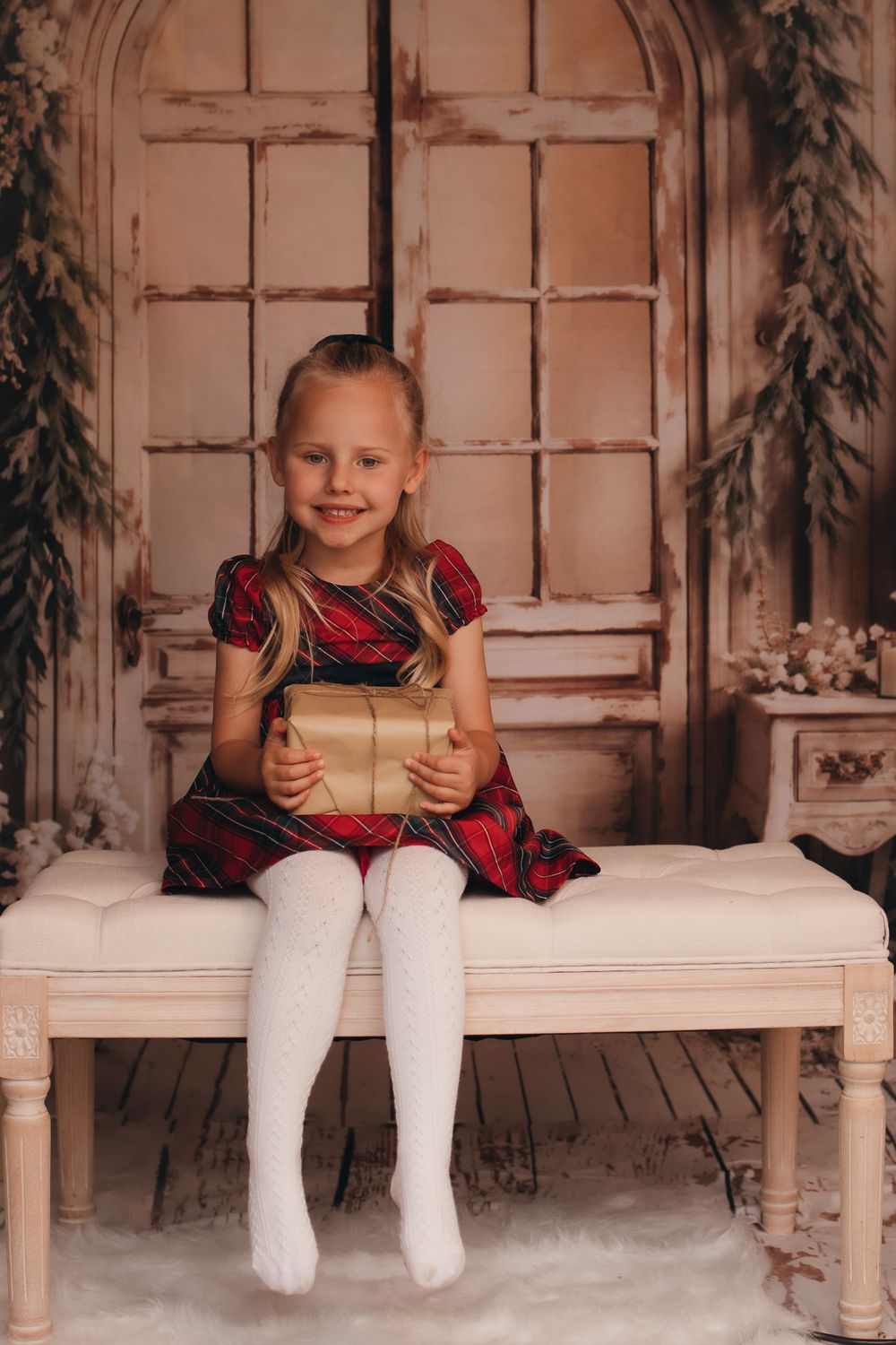 Holiday portrait of child in burgundy dress holding gift on vintage bench setting.
