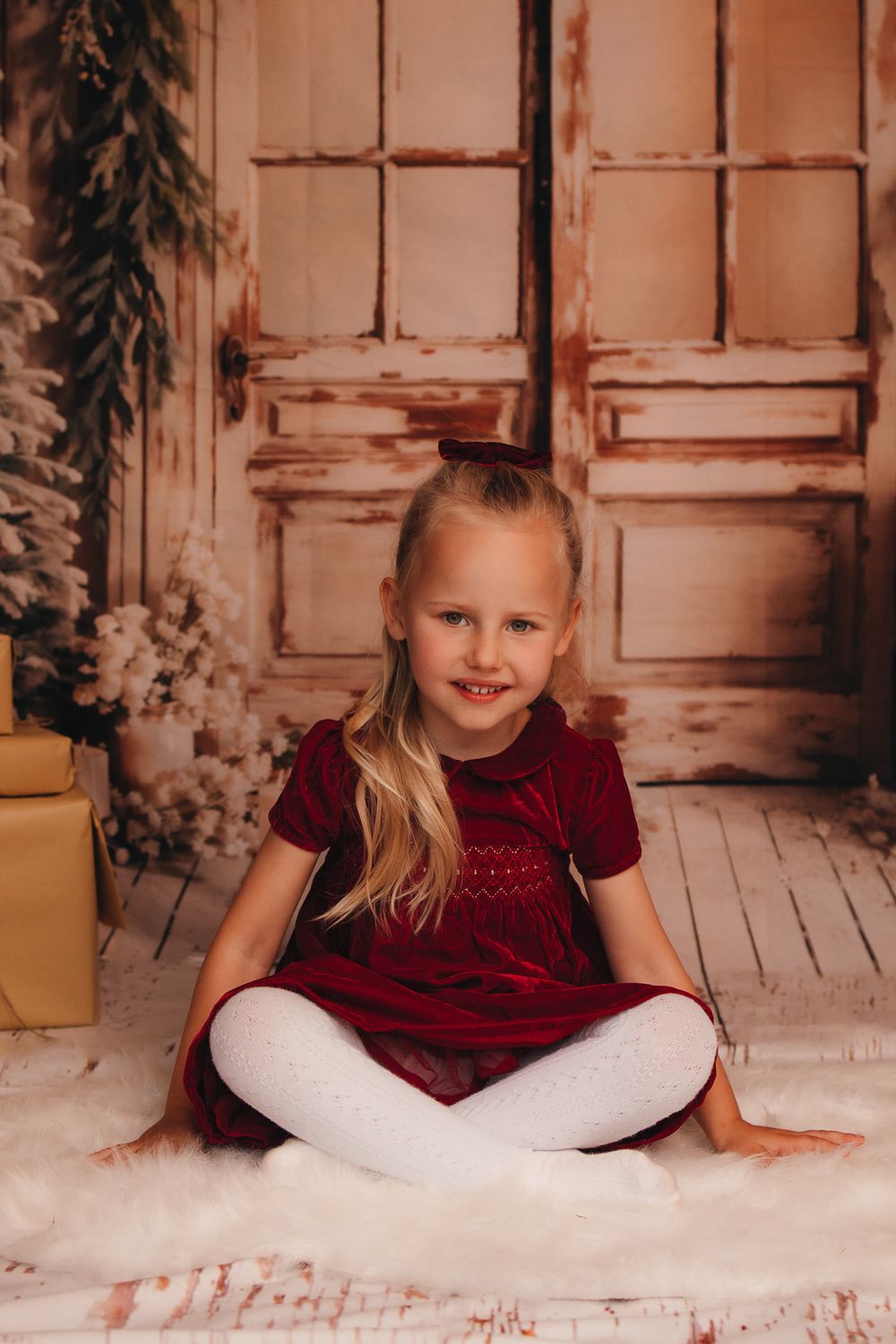 Festive portrait with rustic door backdrop featuring child in velvet dress on fuzzy white rug.