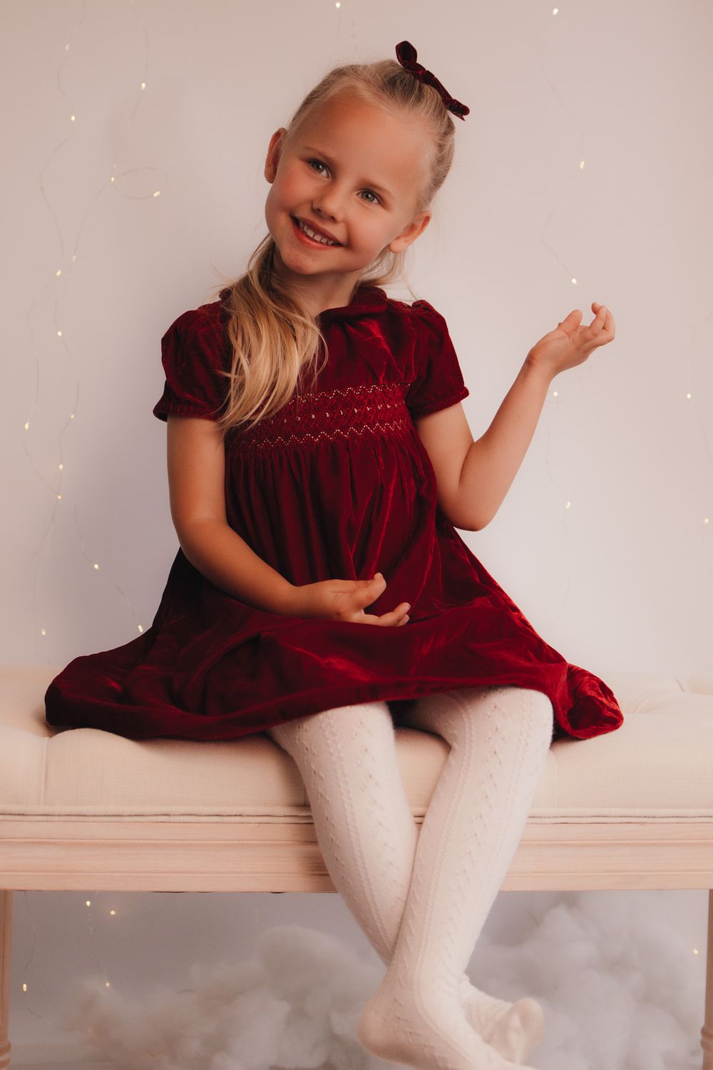 Child in velvet burgundy dress and white tights sitting on bench with sparkly backdrop.