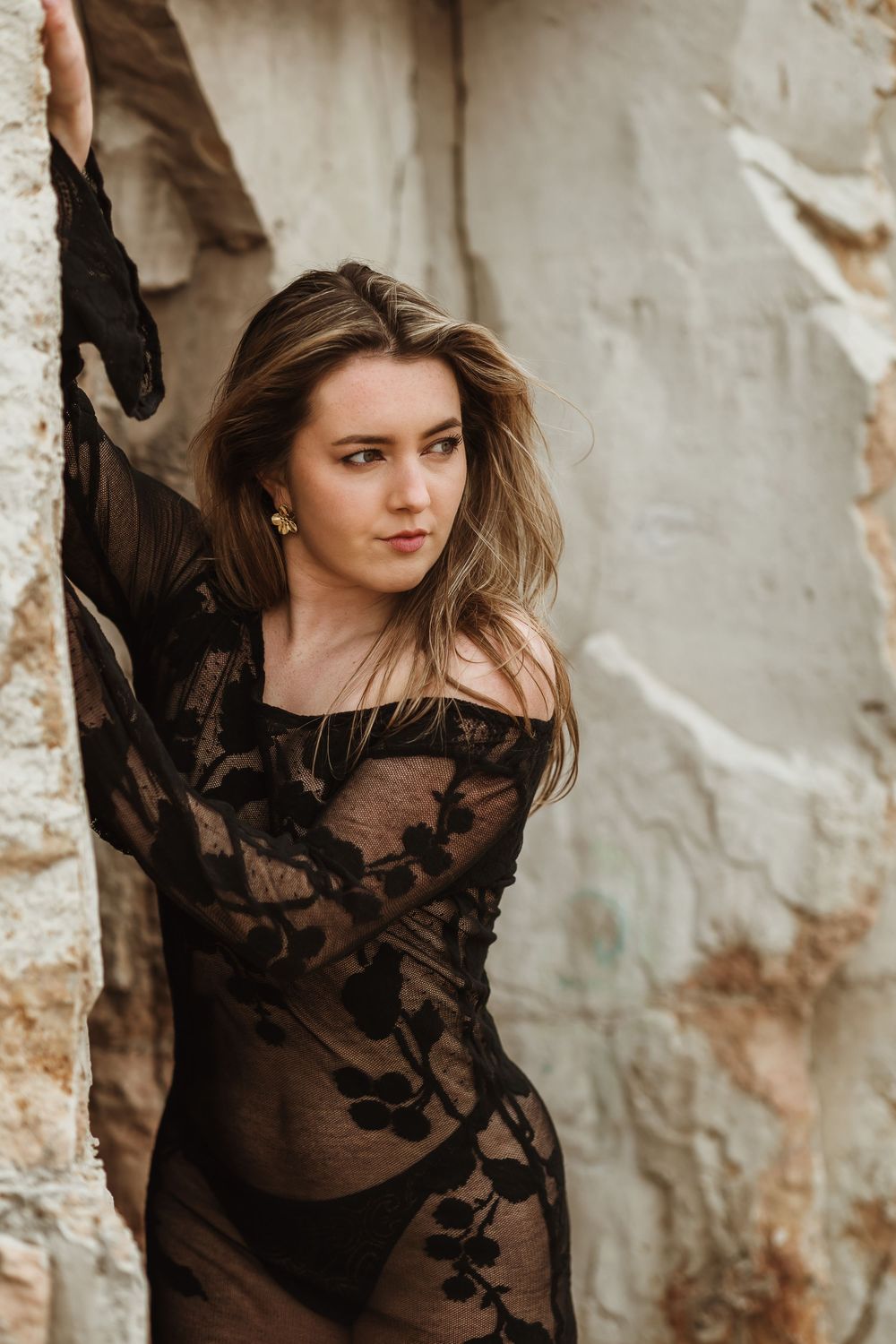Young woman in black lace sheer top posing against rustic stone wall with arm raised, fashion portrait photography.