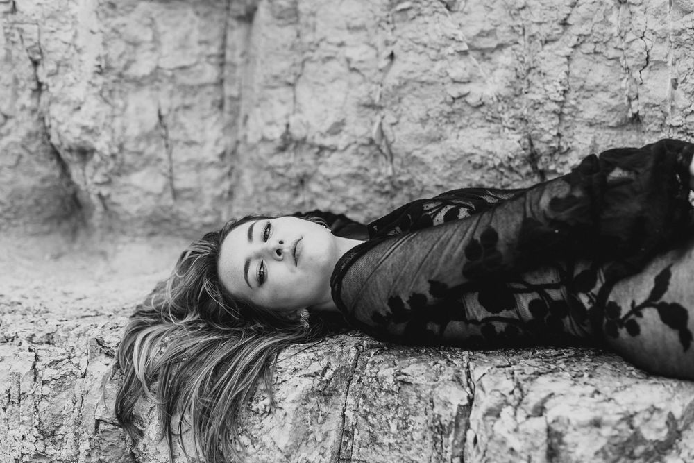 Black and white photo of a woman lying on rocks, wearing a floral lace dress, with her eyes closed.