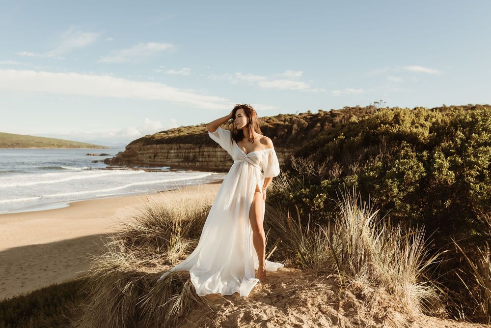Woman in white flowing dress standing on sandy dunes by a scenic beach with rocky headlands and blue sky.
