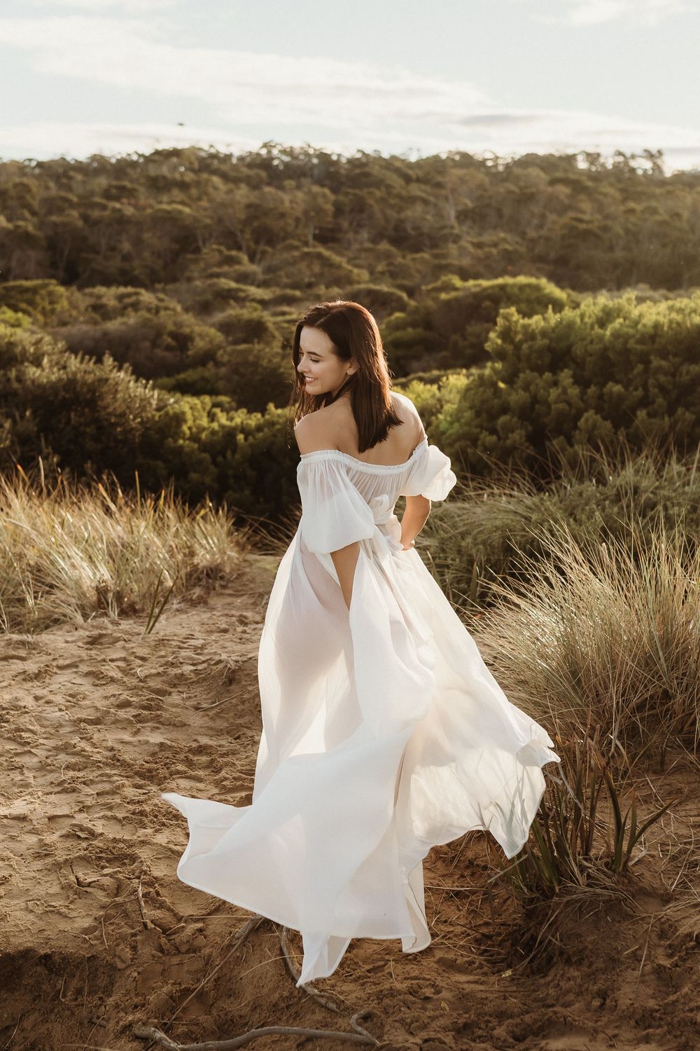 Woman in flowing white off-shoulder dress standing on sandy dunes with lush green bushland backdrop.