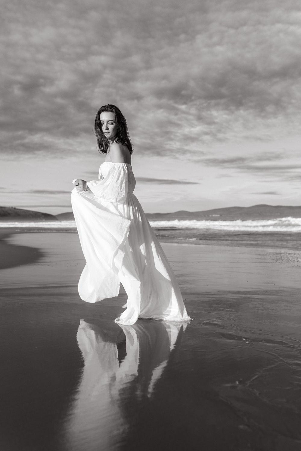 Black and white photo of a woman in a flowing white dress standing on a reflective beach with dramatic cloudy sky.