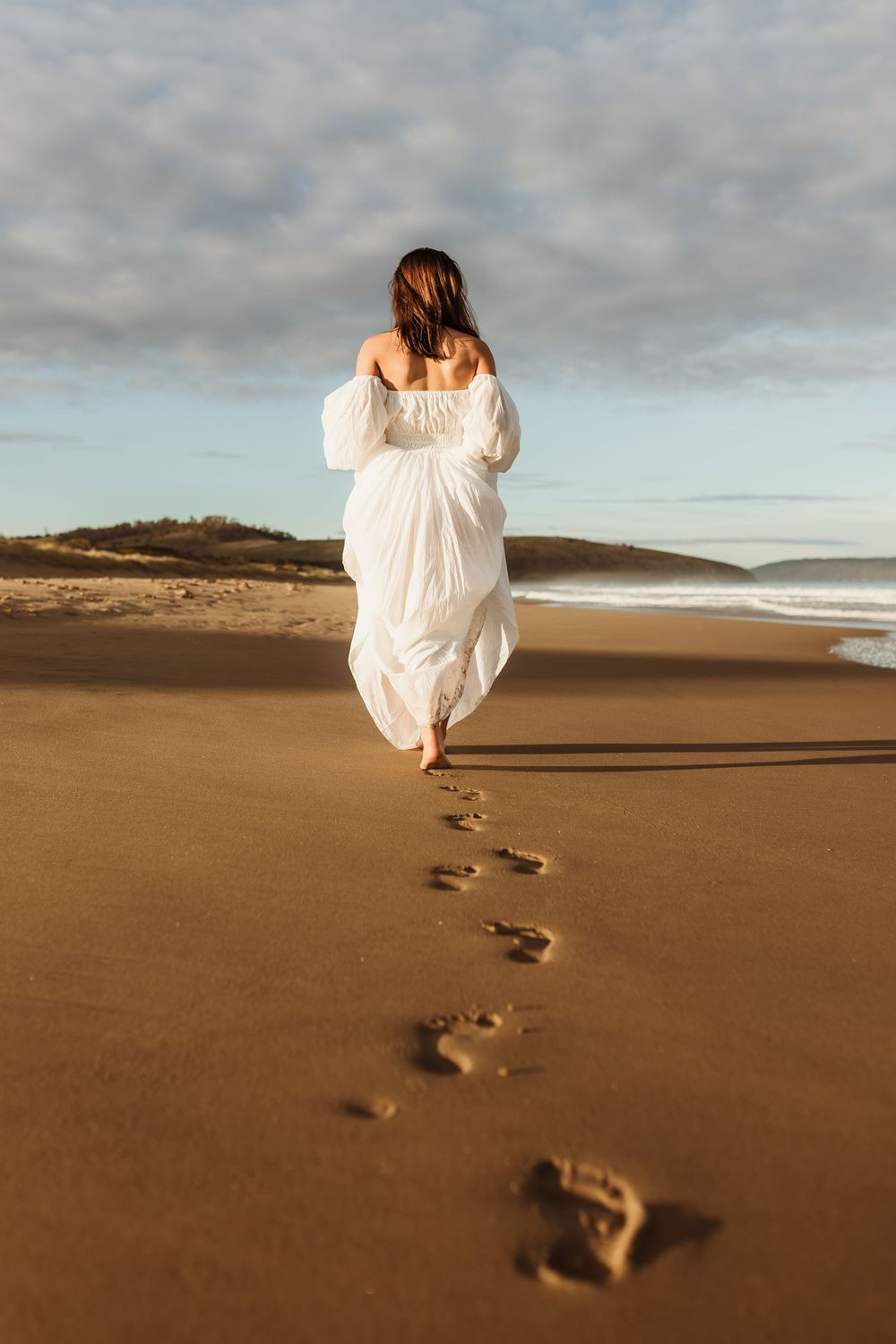 Woman in a white off-shoulder dress walking barefoot on sandy beach leaving footprints at golden hour.