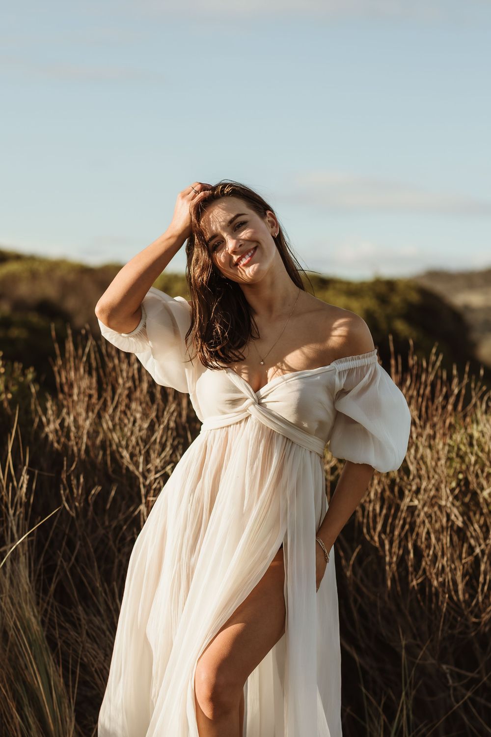 Woman in white off-shoulder dress smiling outdoors among tall grass on a sunny day.