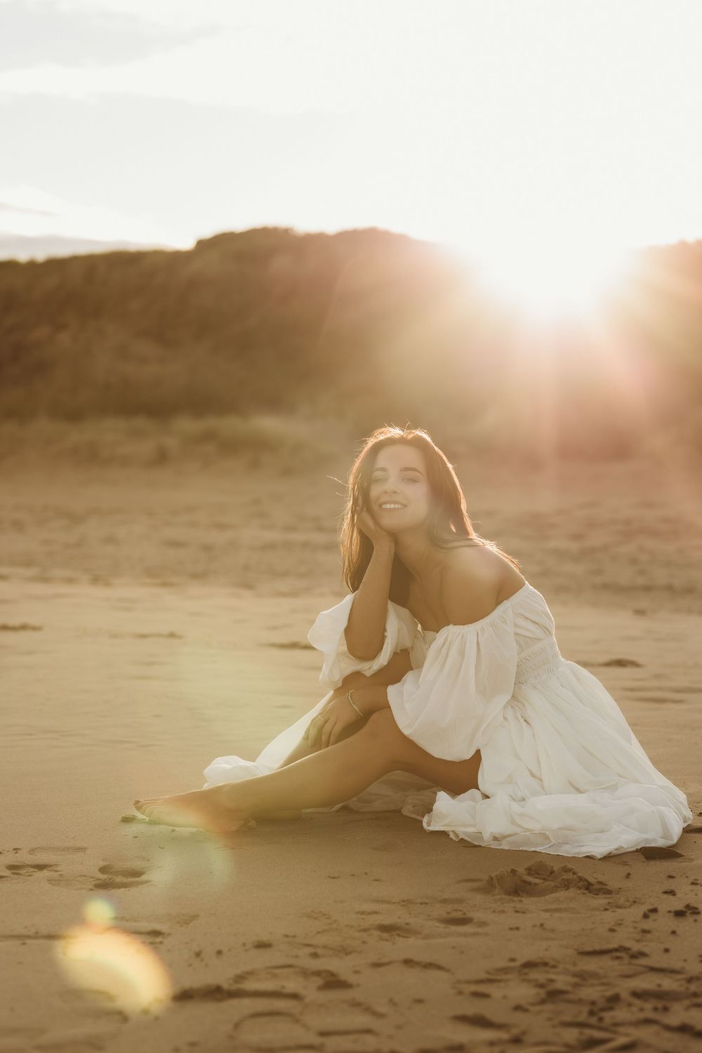 Woman in white off-shoulder dress sitting on sandy beach at golden sunset, smiling warmly with sun flare behind her.