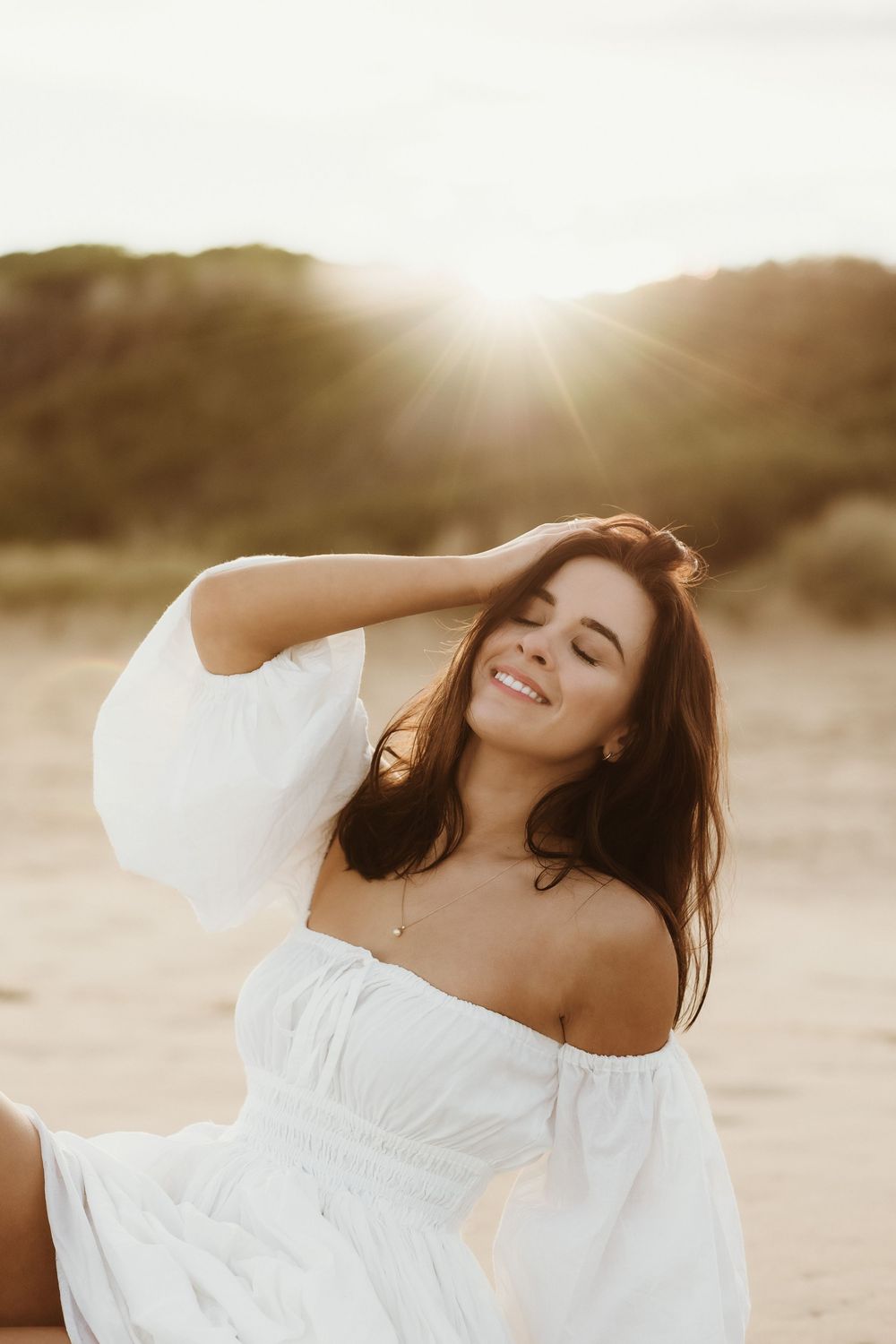 Woman in white off-shoulder dress smiling at sunset on beach, golden hour light glowing behind her.