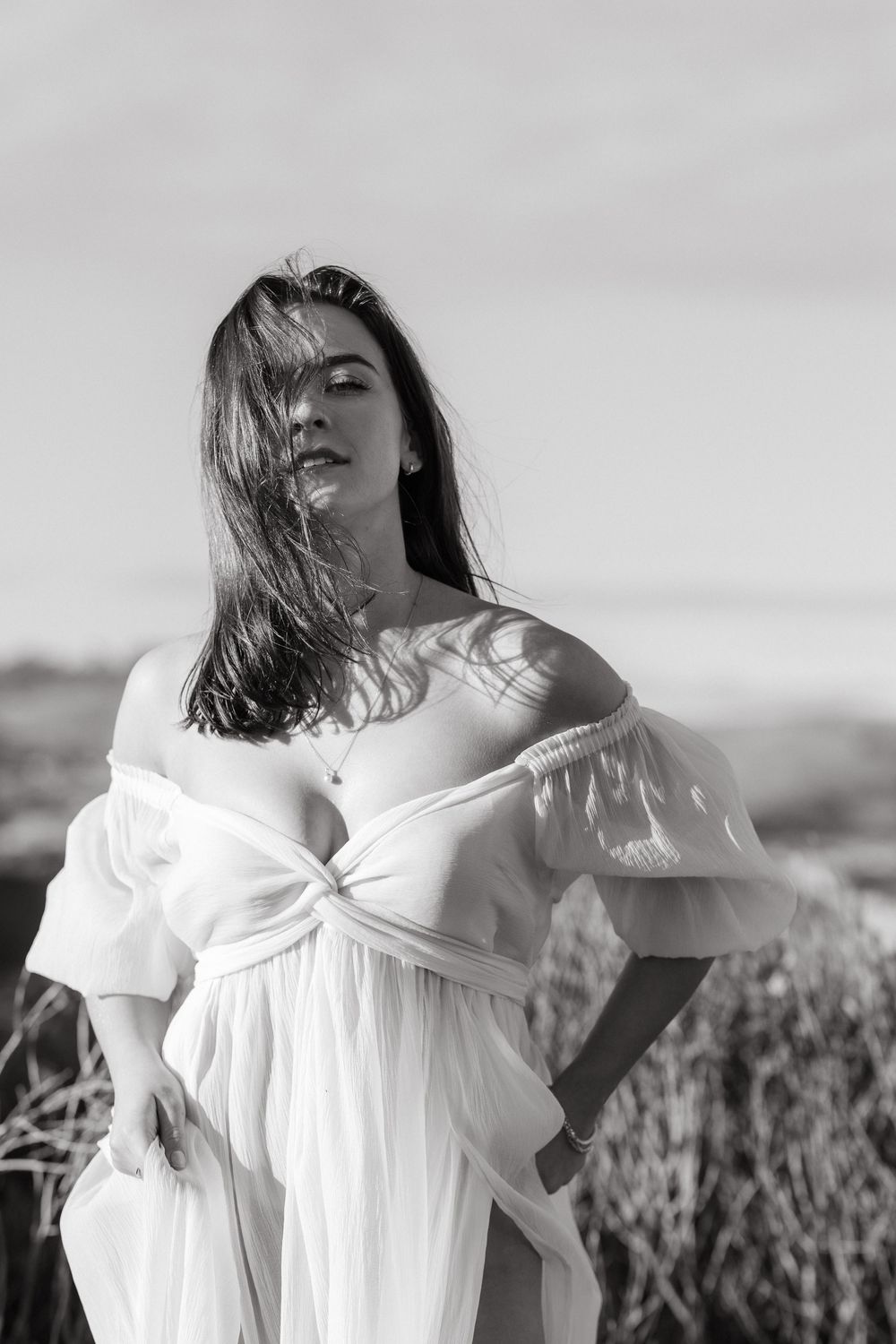 Black and white photo of a woman in a flowing white off-shoulder dress posing outdoors in a natural landscape.