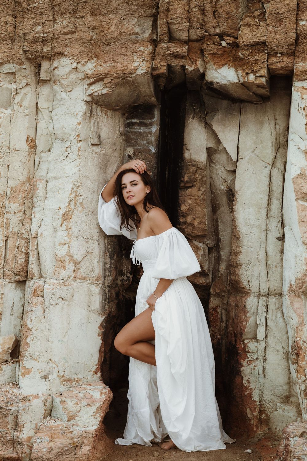 Woman in white off-shoulder dress posing gracefully against rustic rock cave entrance outdoors.