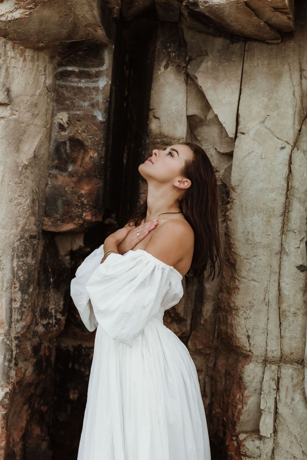 Woman in white off-shoulder dress poses gracefully against rustic stone wall, looking upward with hands on chest.