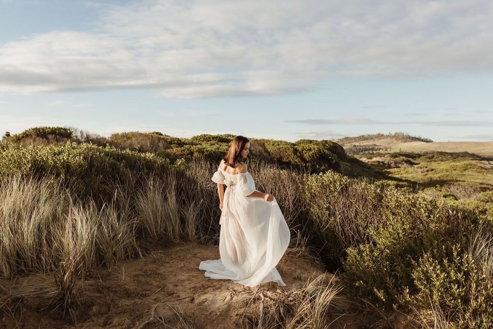 Bride in flowing white gown standing among coastal dunes and wild grasses on a sunny day.