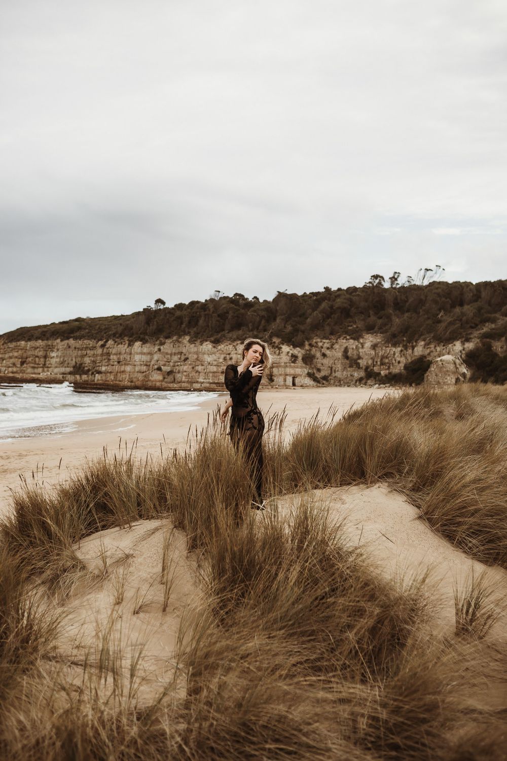 Person poses on sandy dune with coastal cliffs and ocean in background.
