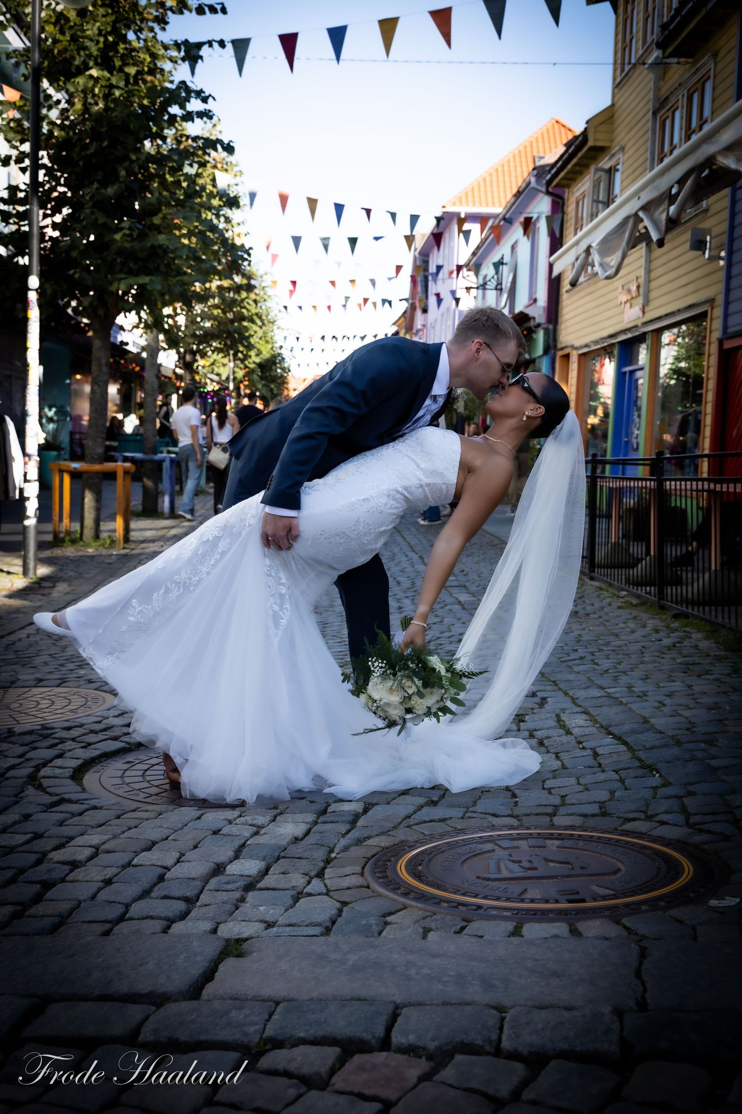 Groom dipping bride for romantic kiss on cobblestone street with colorful bunting flags.