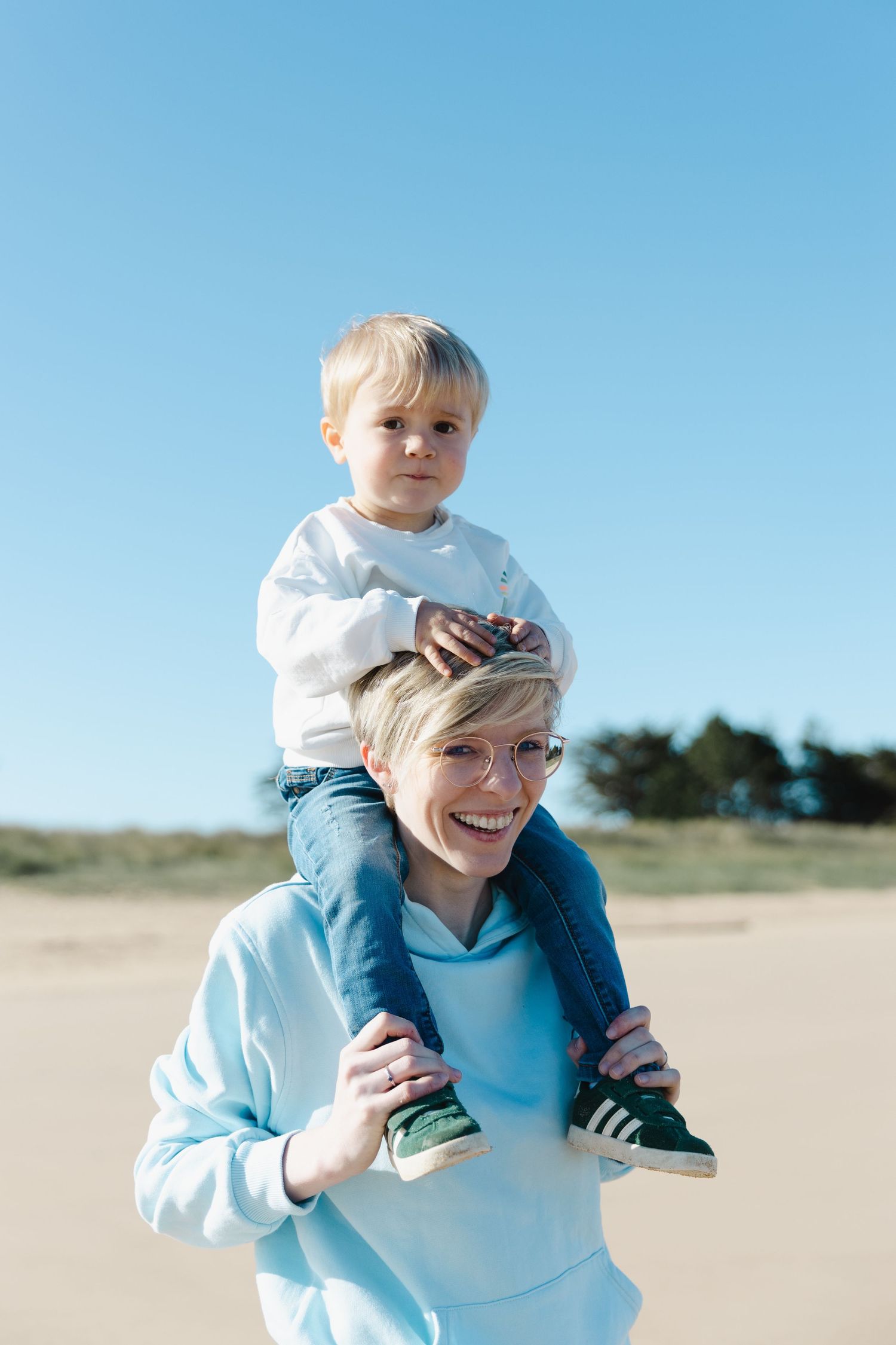 A small child rides on shoulders at a sandy beach on a bright sunny day against clear blue sky.