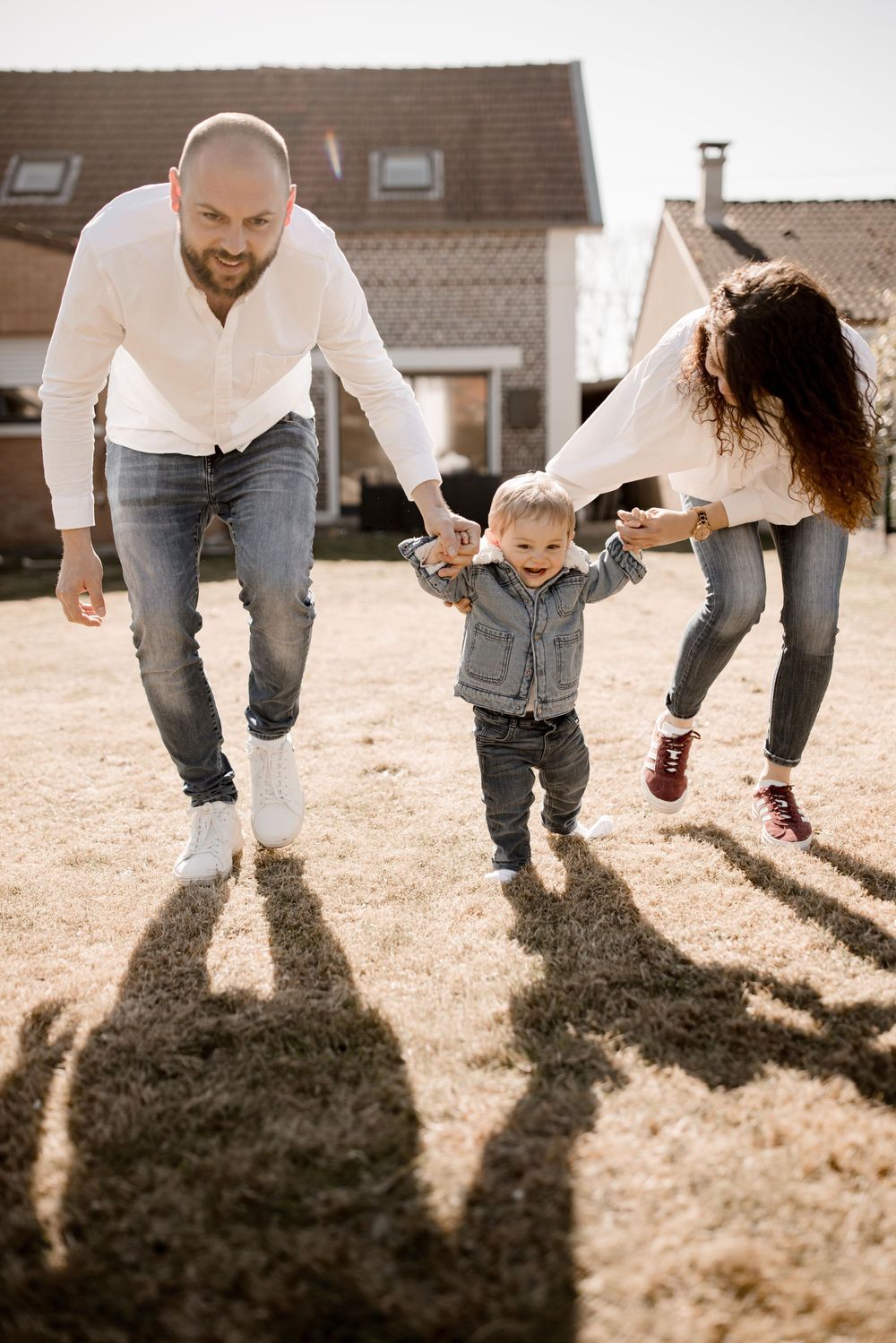 Three people play and dance together outdoors on a sunny day, casting long shadows behind them on the brown grass.