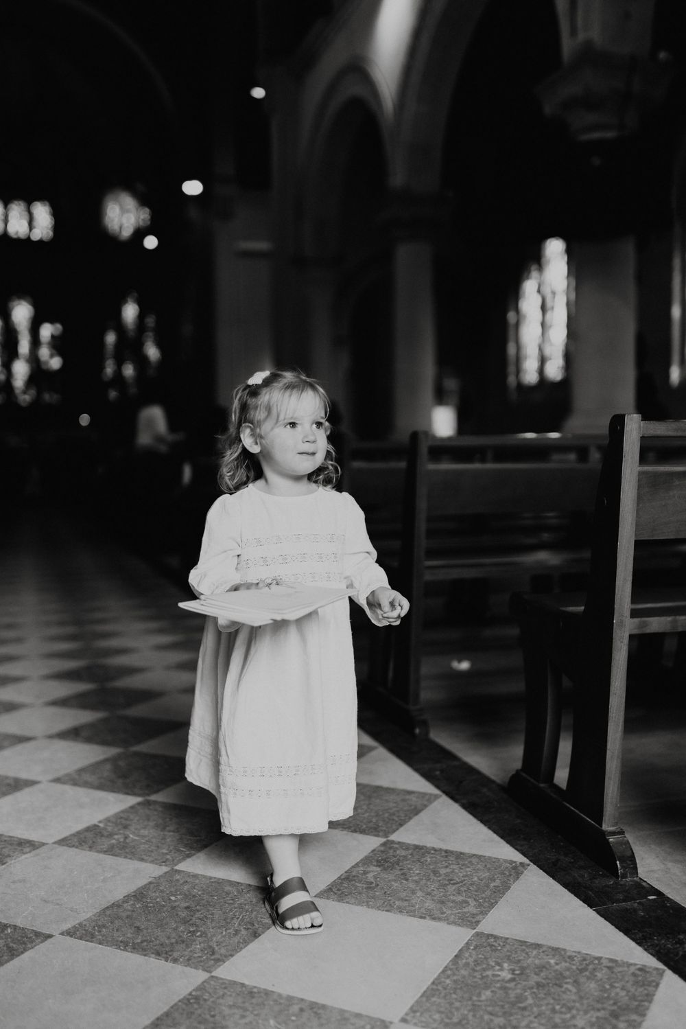 Un petit enfant en robe blanche se tient dans une église au sol carrelé à carreaux lors d'un baptême.