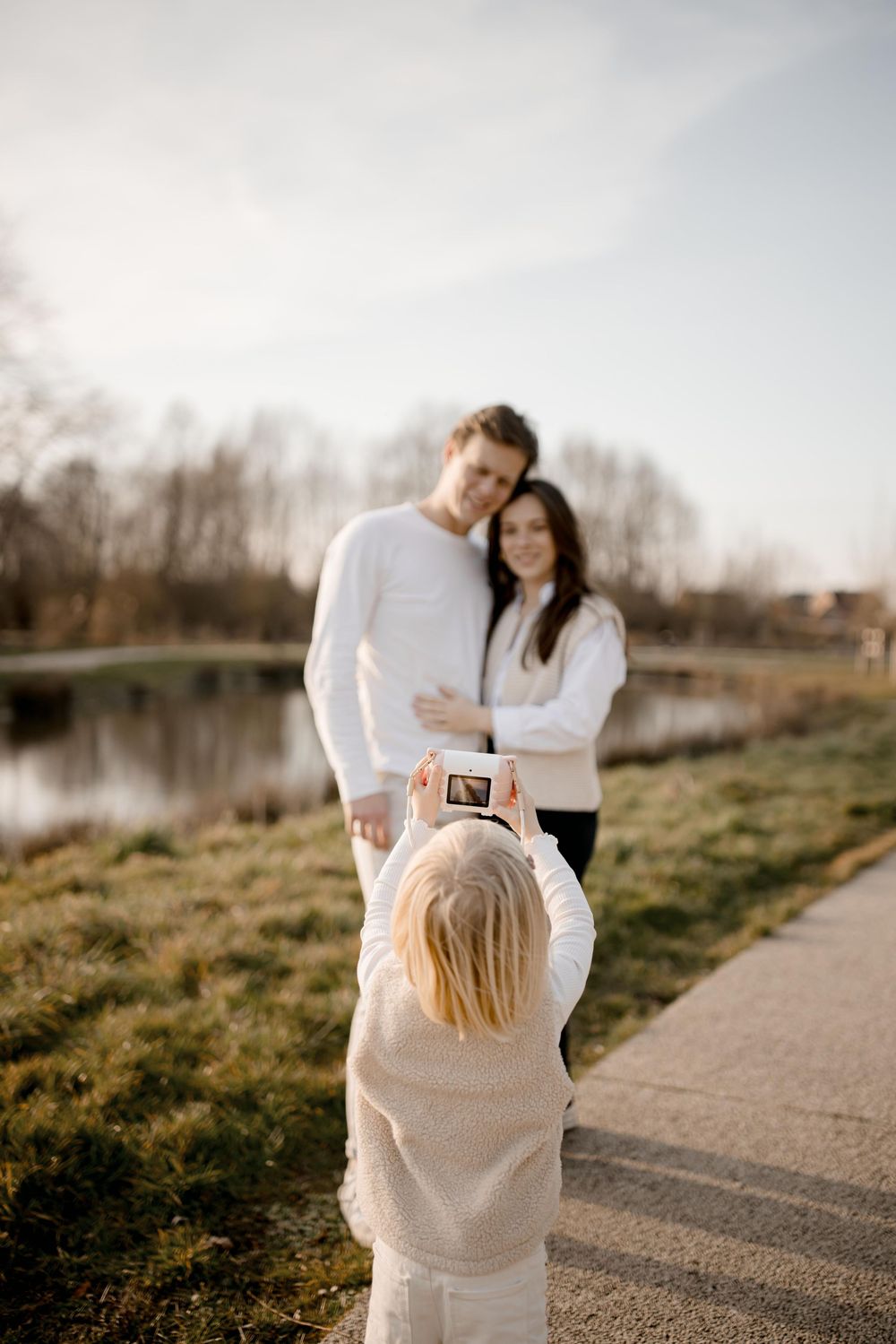 Child takes photo of couple in matching white outfits by a tranquil lake during golden hour.