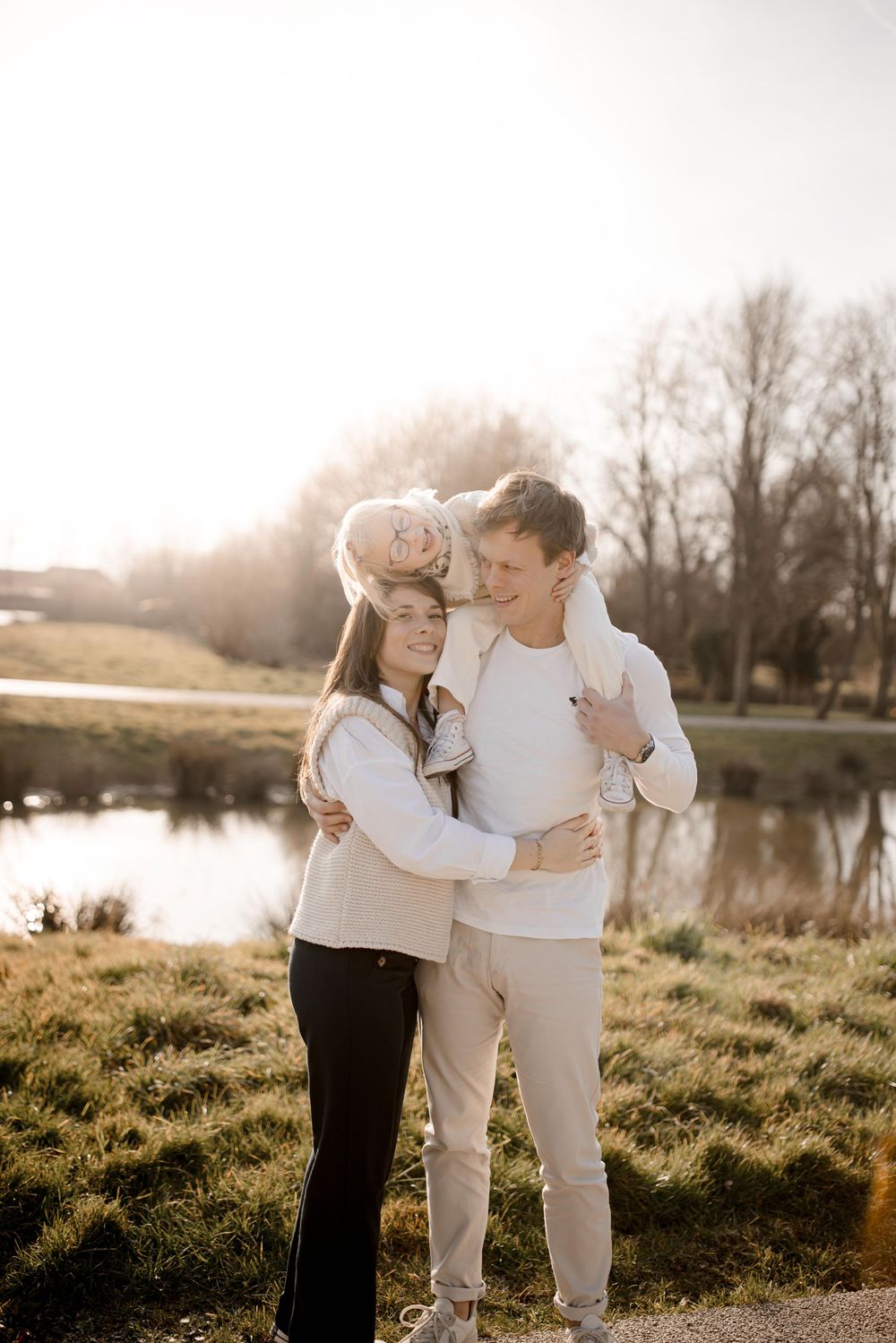 Couple in white sweaters embracing by a lake during golden hour sunset in a tranquil natural setting.