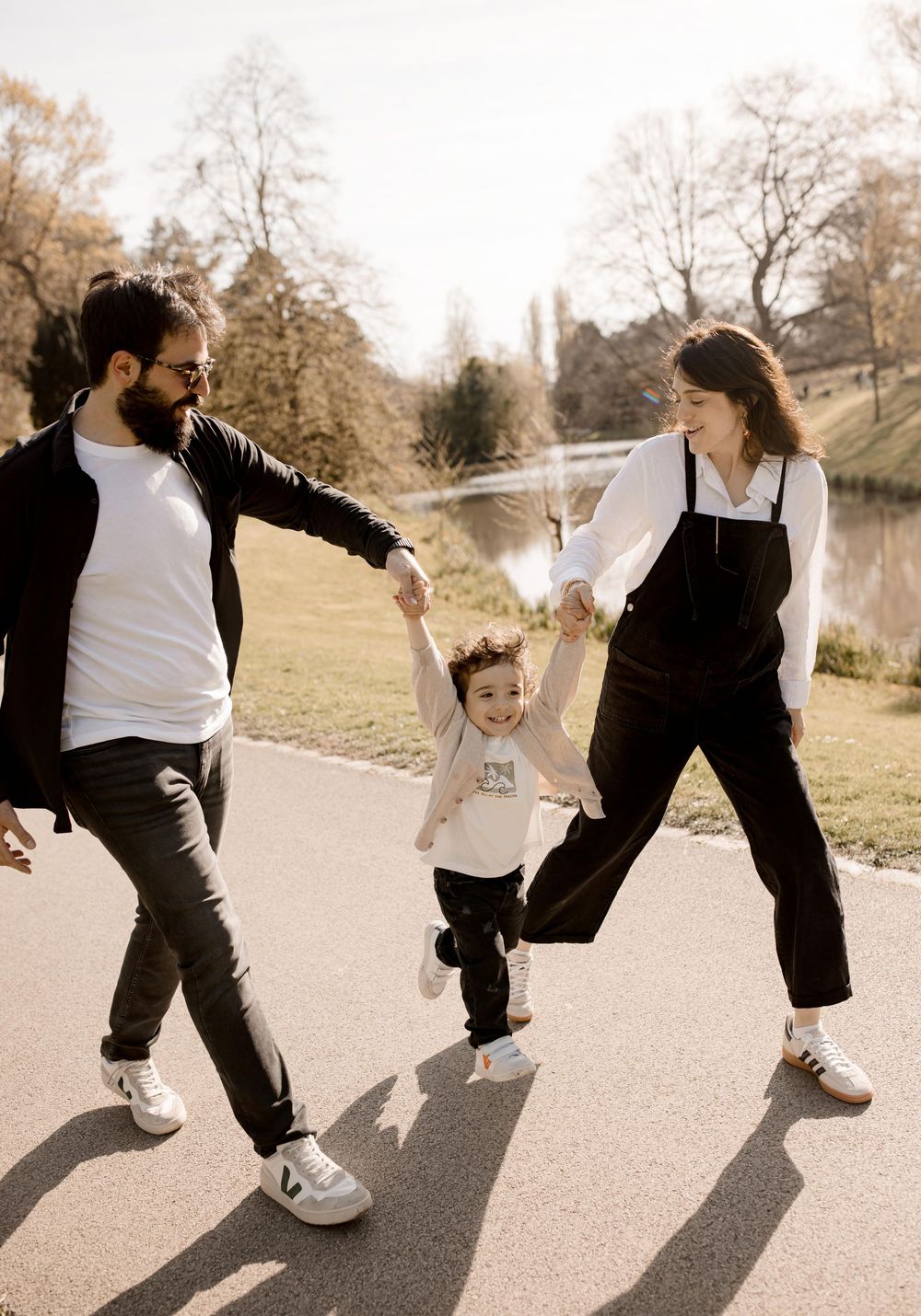 Family in casual outfits swings small child between them while walking on a path near a tranquil pond on sunny day.