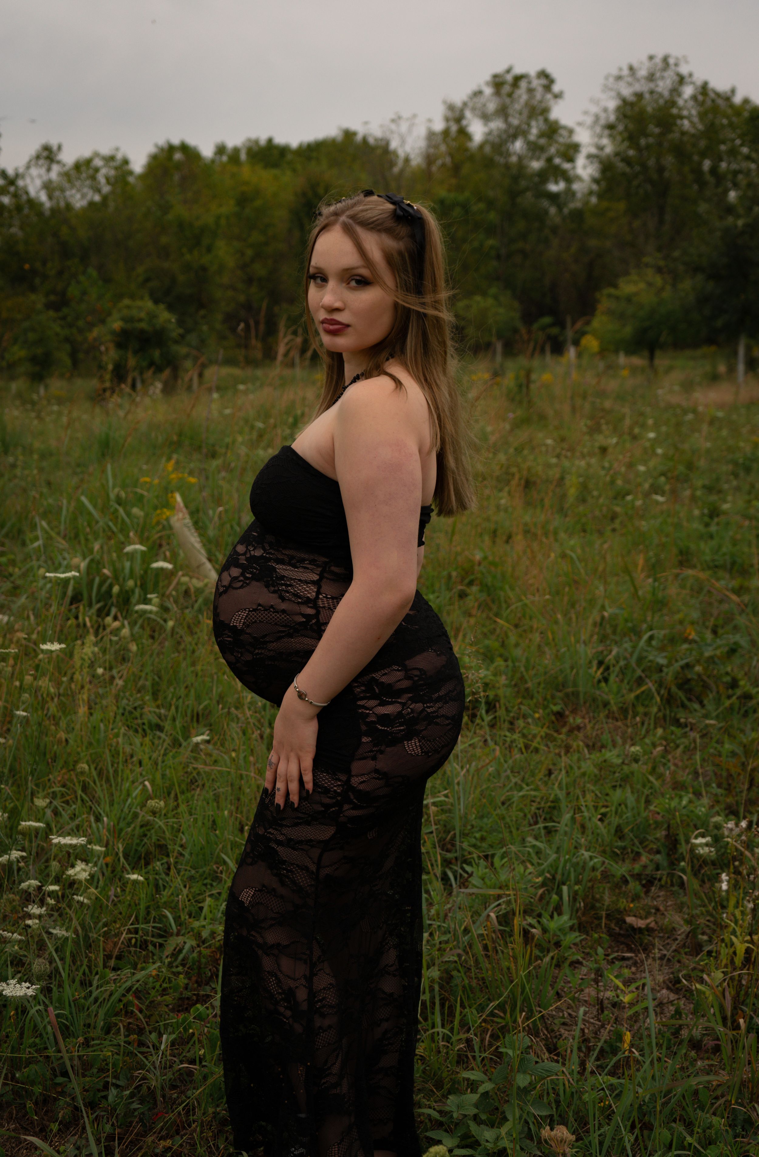Pregnant woman in black lace maternity gown posing in a wildflower field on an overcast day.