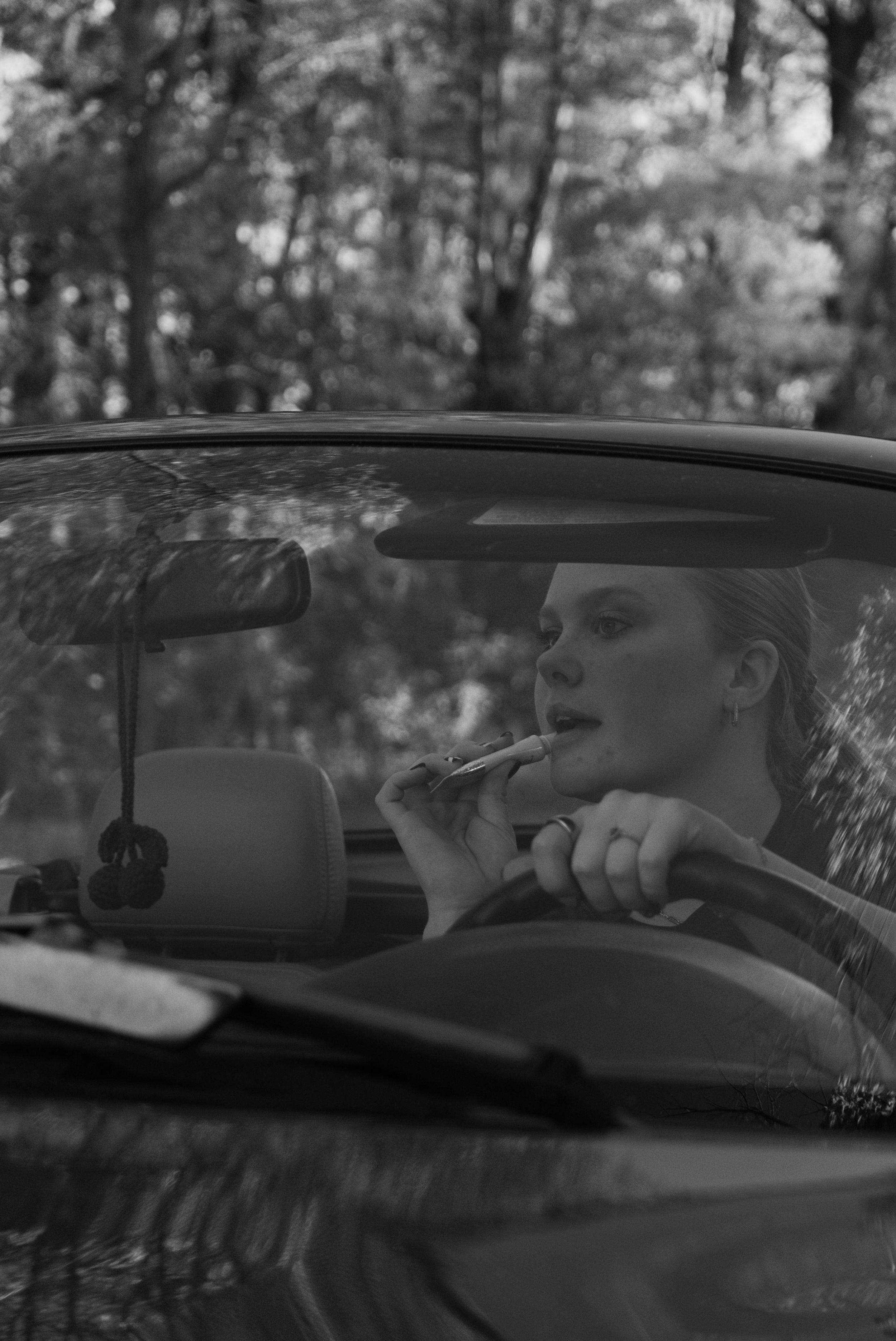 Black and white portrait of a woman applying lipstick while sitting in a car surrounded by trees.