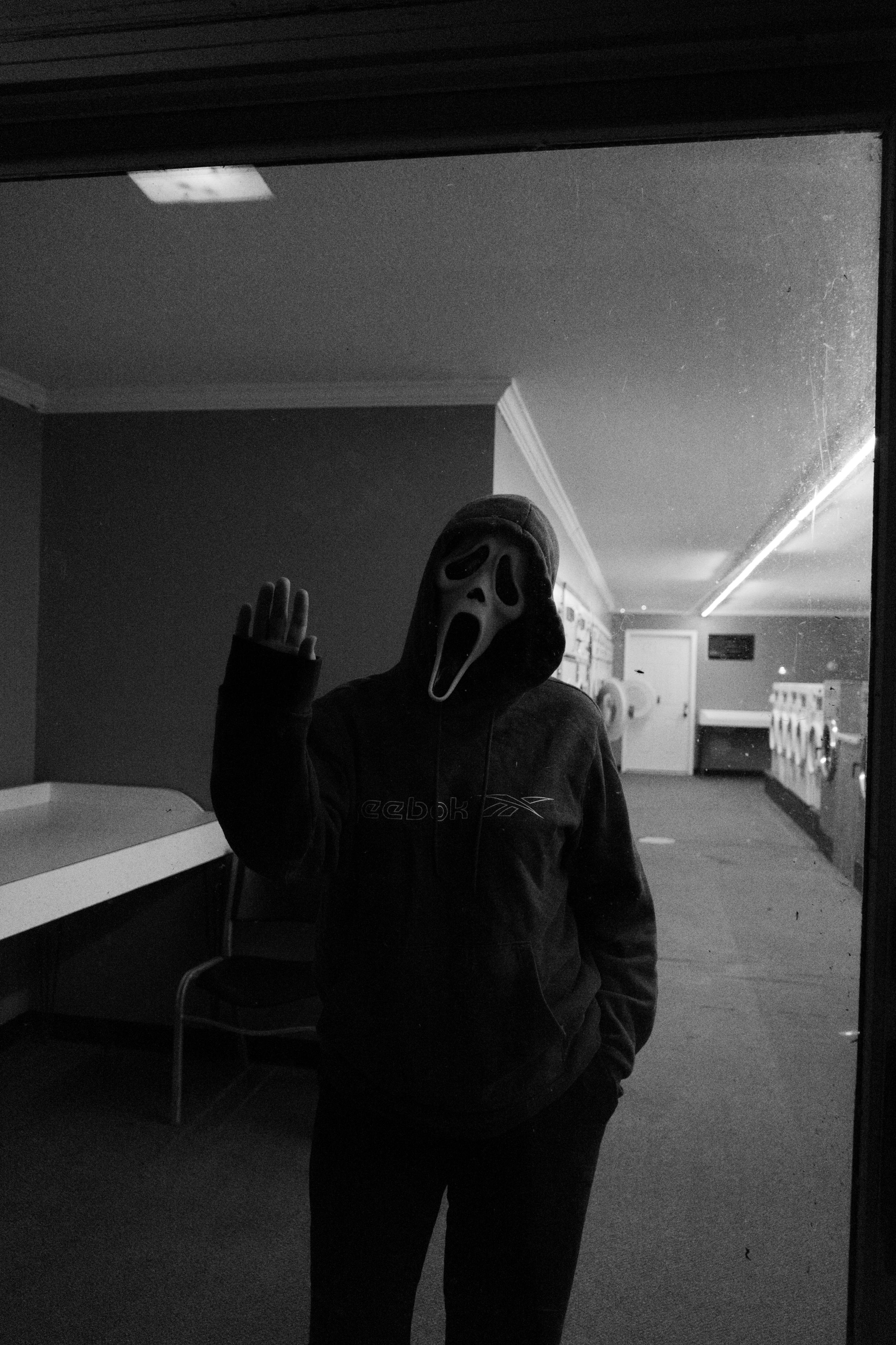 Person in Ghostface Halloween mask waving in a dimly lit laundromat, black and white photo.