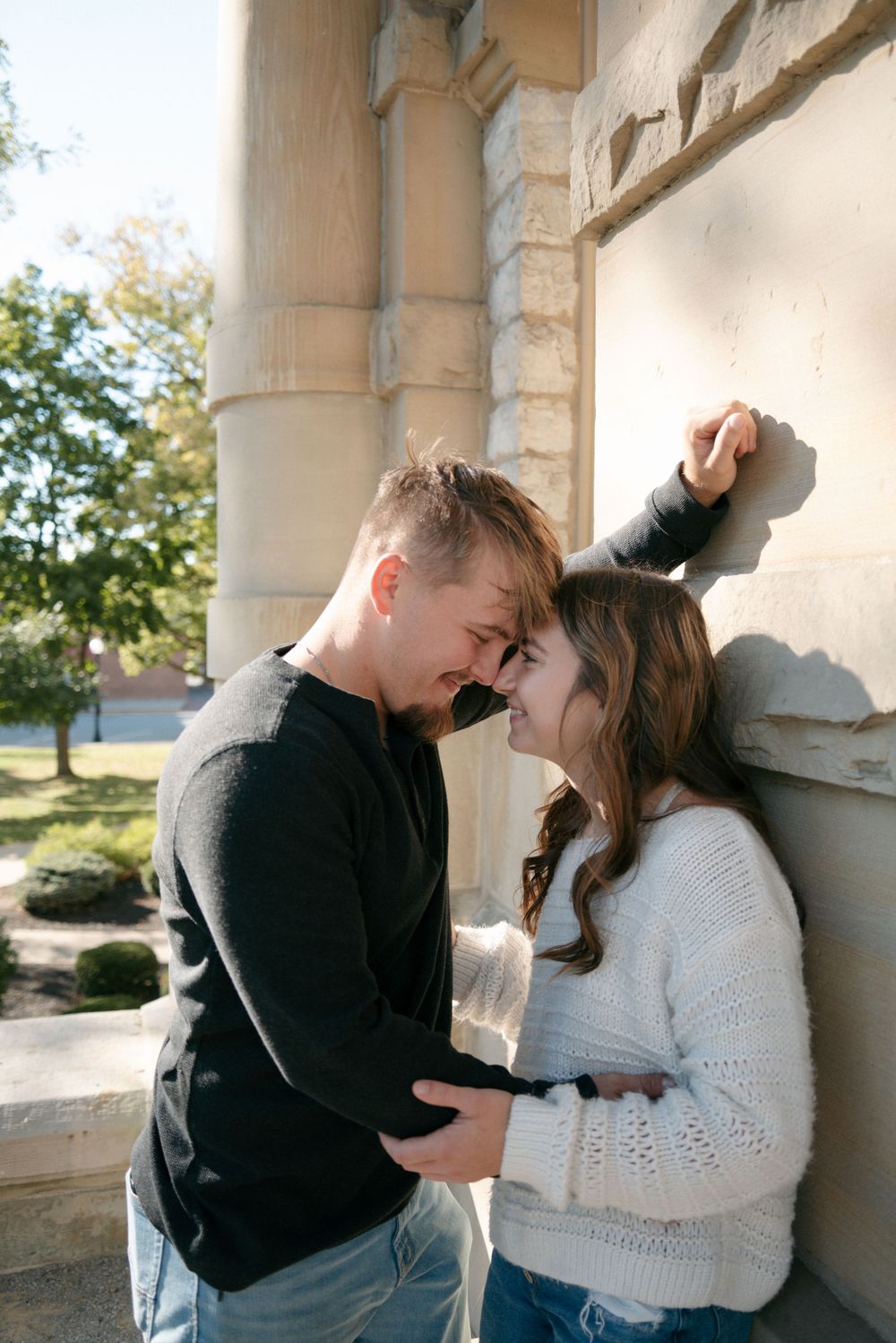 Young couple smiling face-to-face against stone architectural column outdoors in daylight.