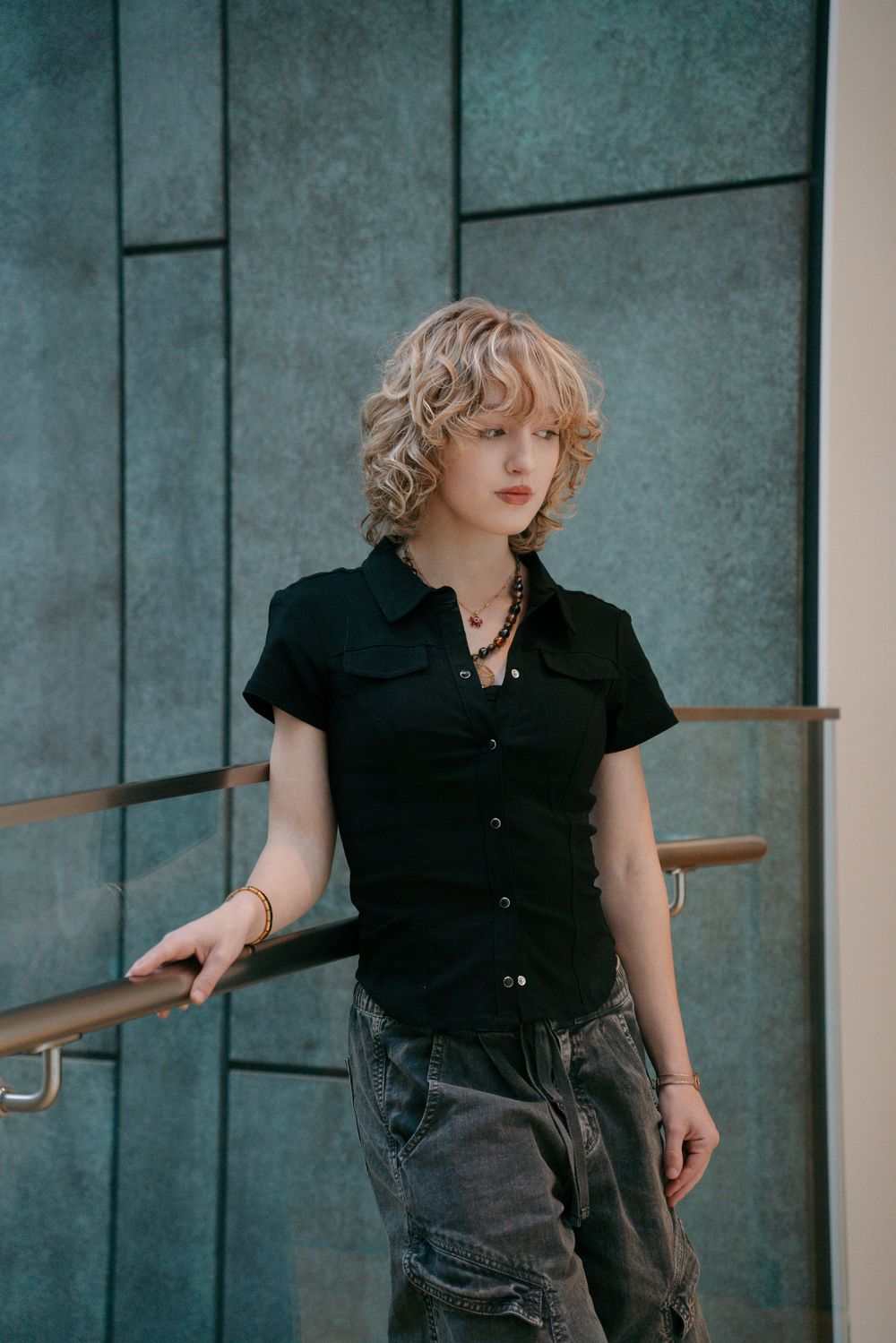Blonde woman in black button-up shirt and gray jeans leaning on railing against teal wall.