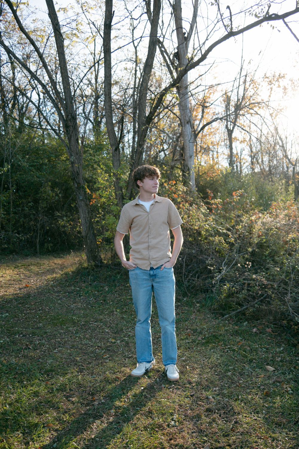 Young man in beige shirt and jeans standing outdoors among autumn trees with hands in pockets.